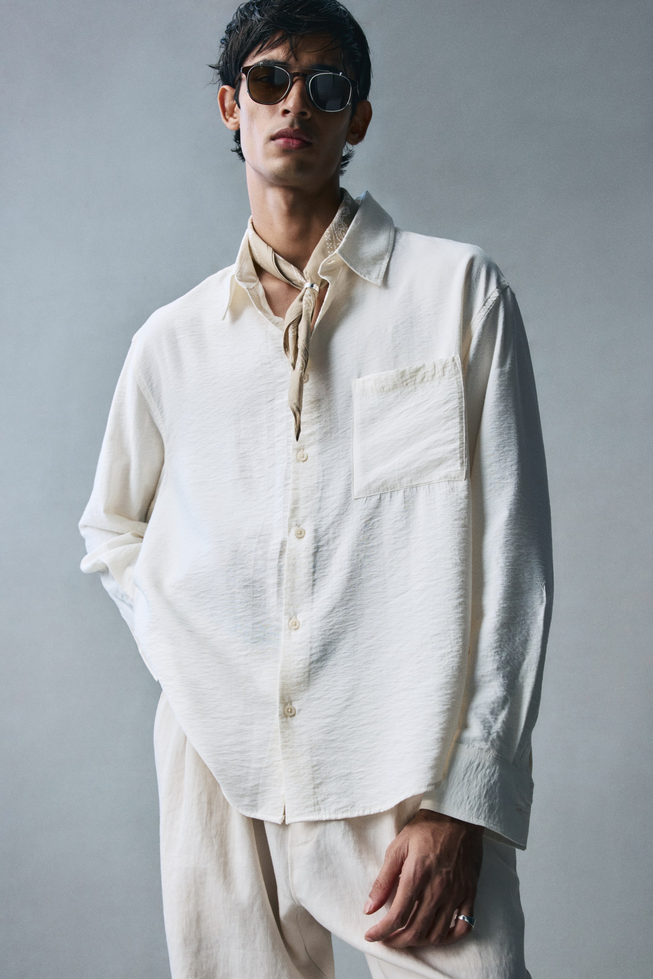 View larger image: The model is wearing a light cream-coloured shirt in a crinkled weave, featuring a relaxed fit, a collar, a buttoned front, and a single patch chest pocket. The long-sleeved shirt is styled untucked with matching light-coloured trousers and a patterned neck scarf.