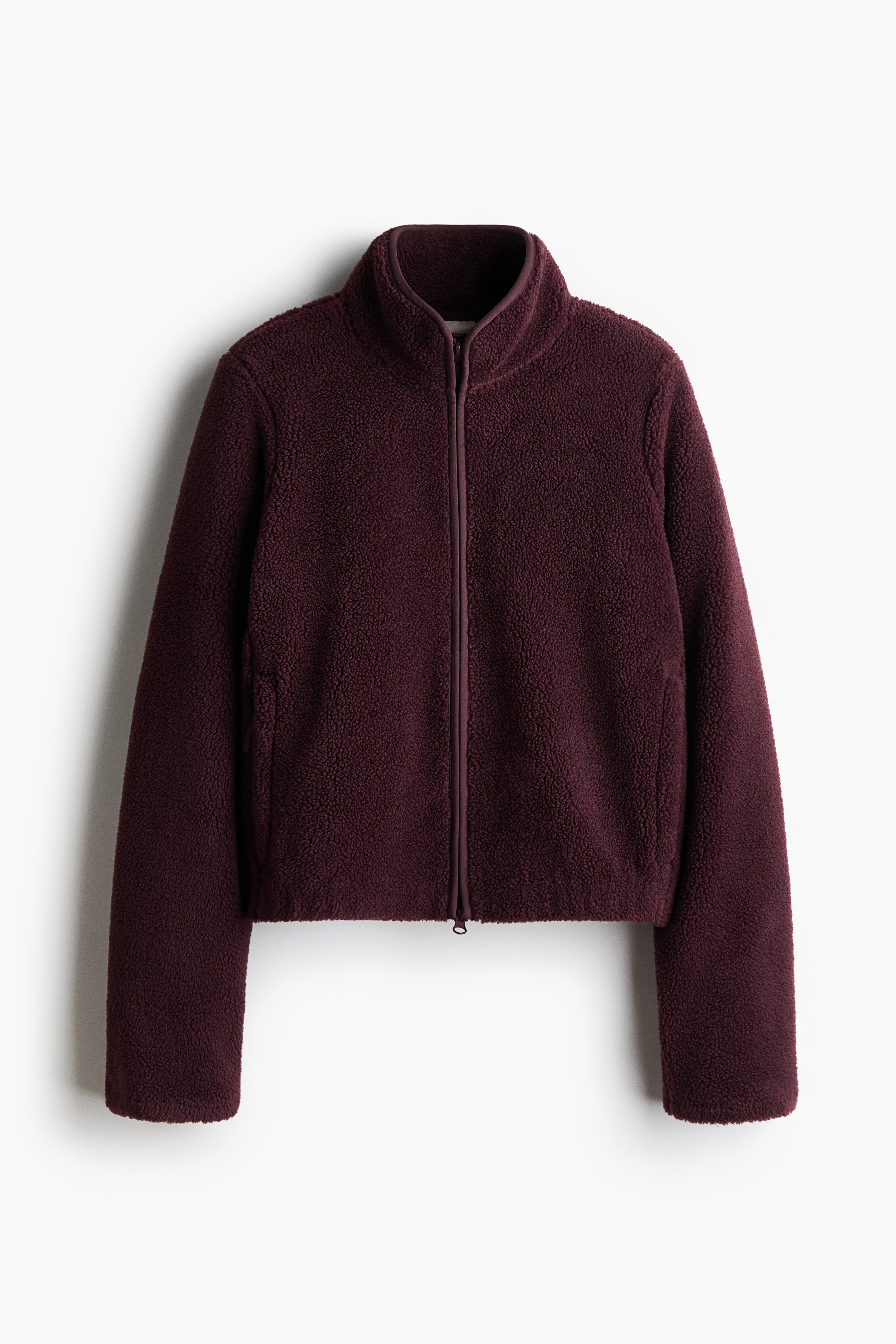A dark burgundy fleece jacket with a high collar and front zipper is worn by the model, who is holding the front opening with one hand. The jacket is styled with matching mauve leggings visible at the waist.