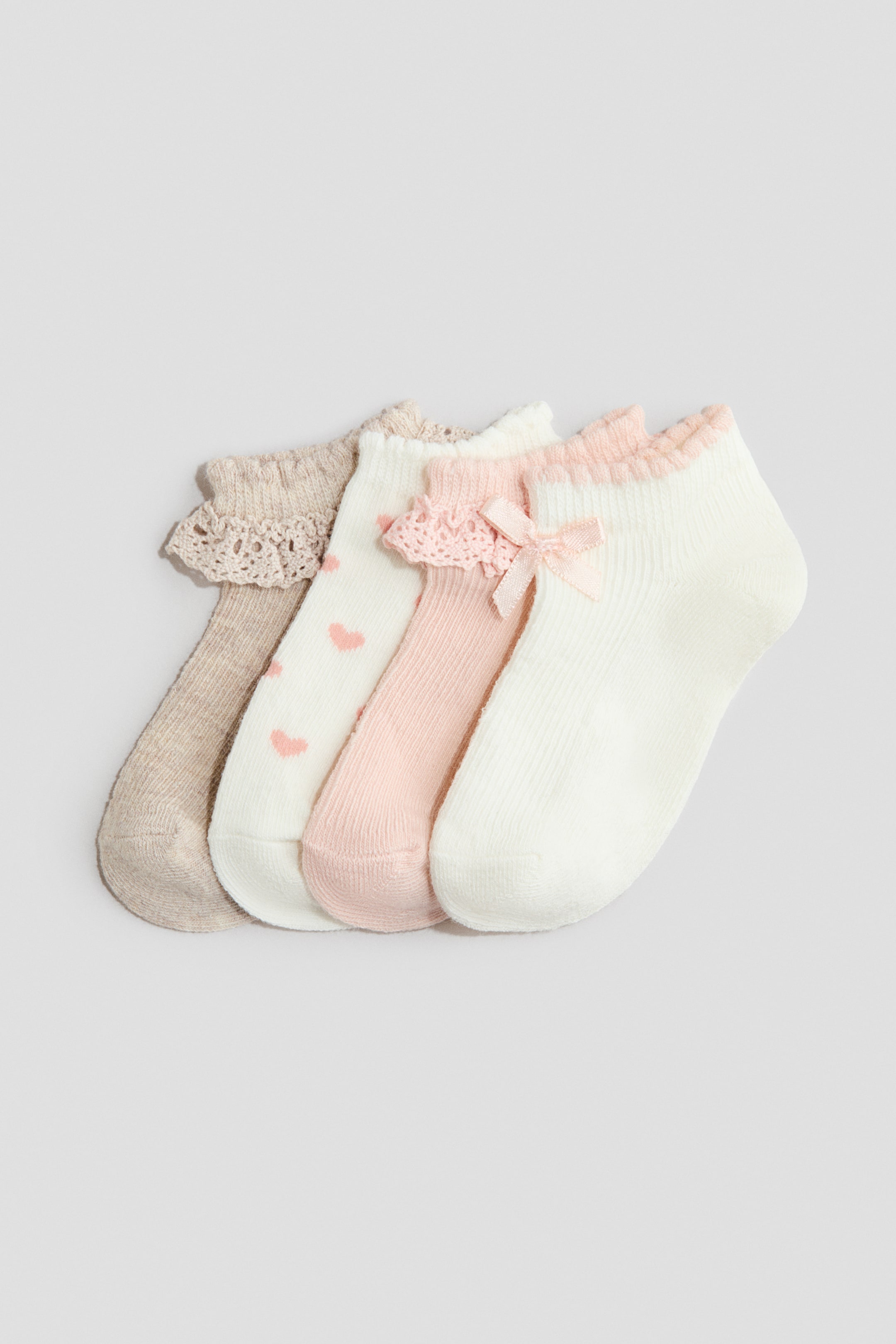 Visa större bild: Fyra skaftlösa strumpor i en mjuk, finstickad bomullsblandning visas framifrån, var och en med en elastisk topp med dekorativa detaljer. Designen inkluderar beige med virkad spets, vit med rosa hjärtan, rosa med virkad spets och en satinrosett, samt vit med en rynkad rosa ribbad kant.