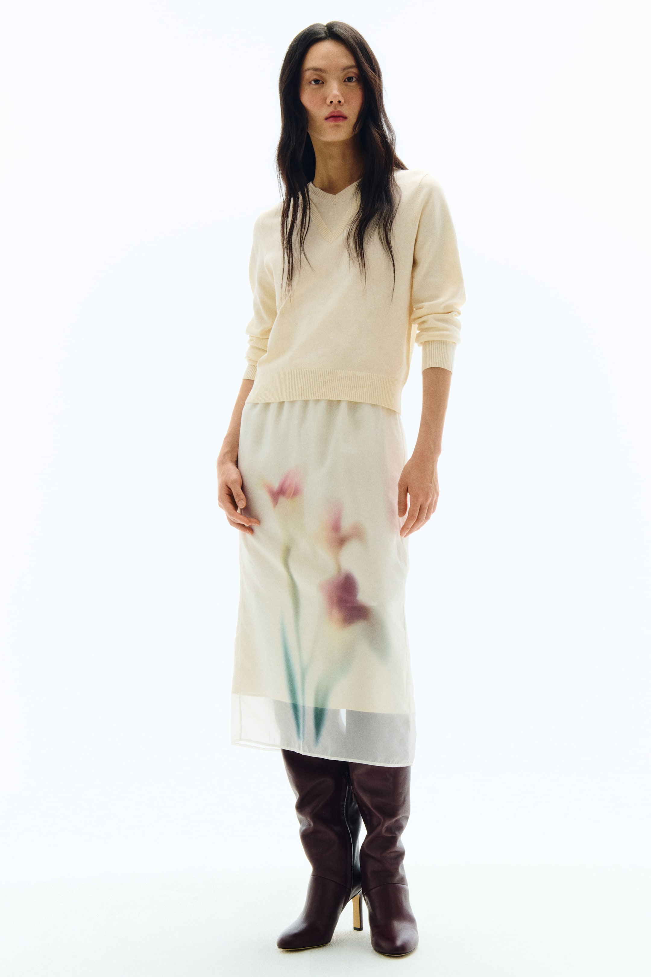 拡大画像を表示: ライトクリームカラーのVネックセーターは、袖口と裾にリブ編みのディテールが施されており、シアーな淡色のミディ丈スカートとスタイリングされています。スカートは、ピンク・パープル・グリーンのぼかしたフローラル柄が、透け感のある生地から不透明なライニングの上に重なるようにあしらわれており、ポインテッドトゥとブロックヒールが特徴のダークブラウンのニーハイブーツとコーディネートされています。