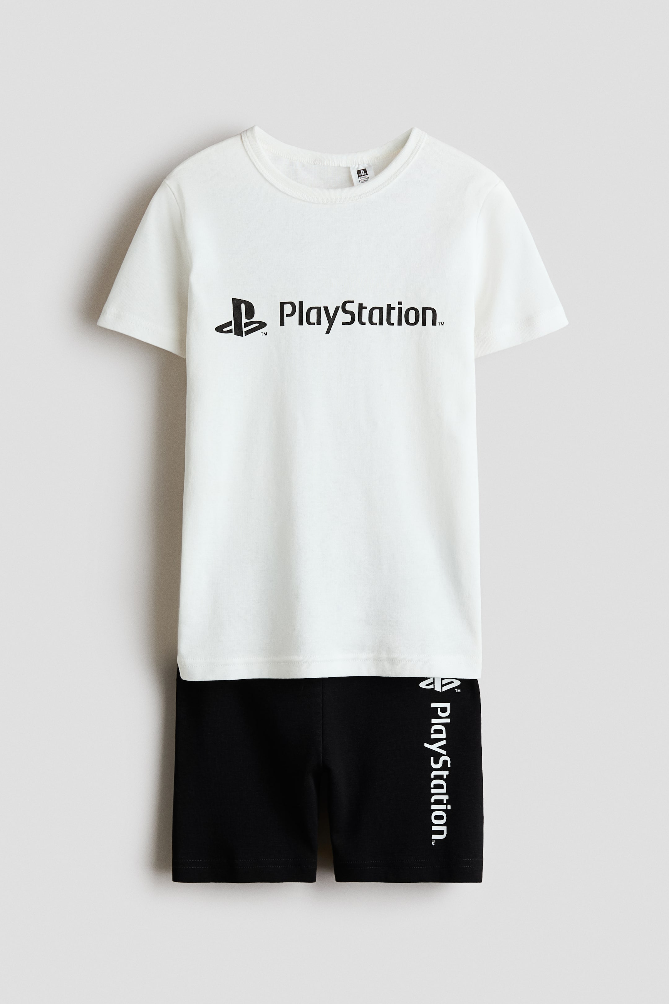 Voir une image plus grande: Un ensemble de pyjamas en jersey de coton doux est présenté, comprenant un t-shirt blanc avec un col rond bordé de côtes et un logo PlayStation noir imprimé sur le devant. Un short noir accompagne le t-shirt, avec une taille élastiquée recouverte et un logo PlayStation blanc vertical imprimé sur la jambe gauche.