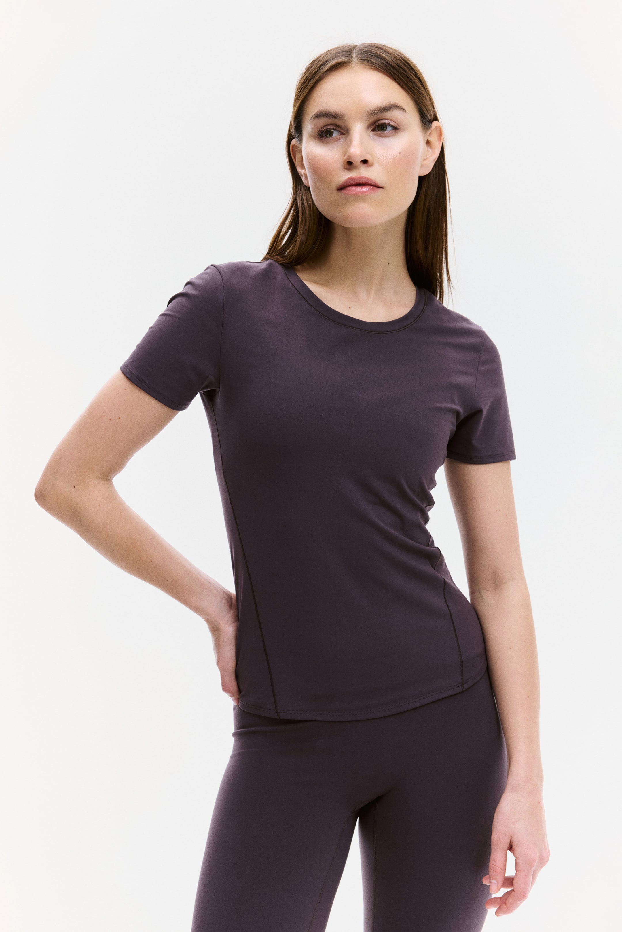 Ver imagem maior: Uma t-shirt de manga curta em roxo-acinzentado escuro com gola redonda está a ser usada, apresentando um design ajustado e costuras laterais visíveis. Está a ser combinada com leggings a condizer.