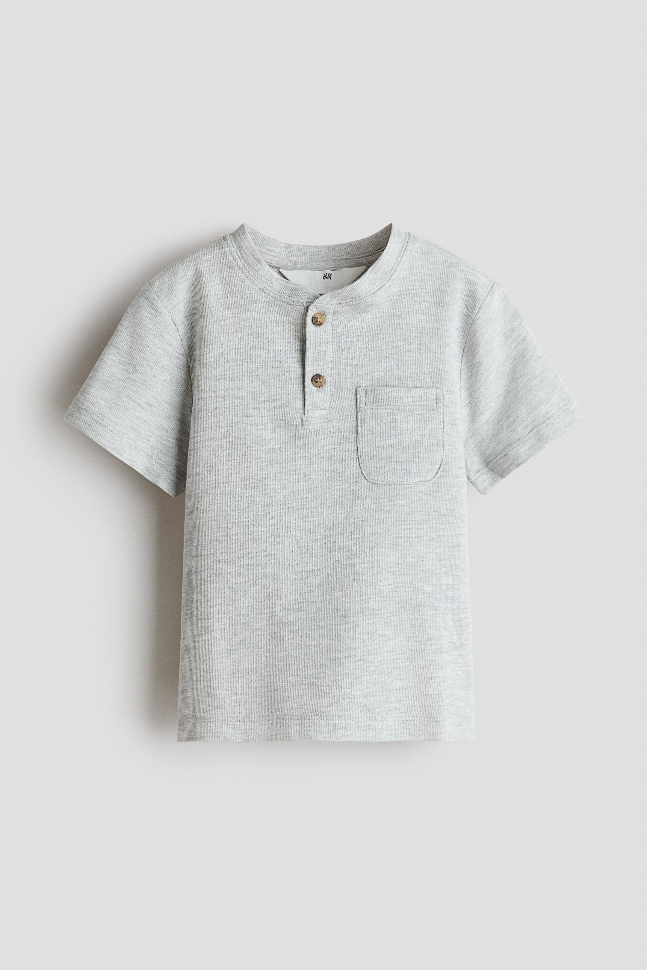 View larger image: A light grey short-sleeved henley shirt is visible from the front, featuring a round neckline with a two-button placket and brown buttons. It also displays a patch pocket on the left chest and showcases a fine textured knit fabric.