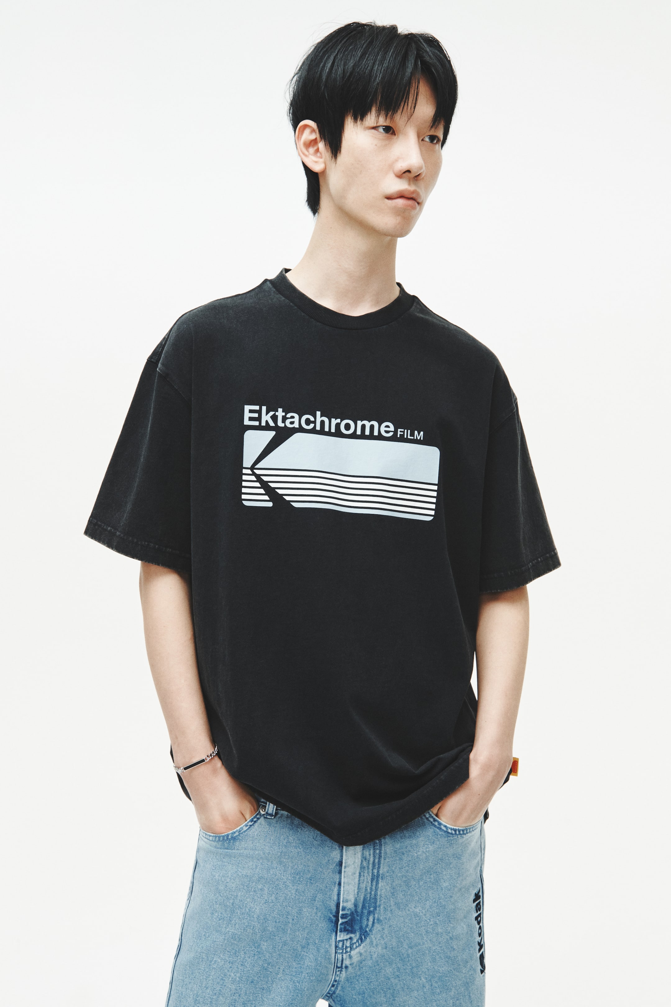 拡大画像を表示: 黒のオーバーサイズTシャツが正面から紹介されています。胸部には大きな長方形のグラフィックがあり、白い文字で「Ektachrome FILM」と表示されています。グラフィックにはスタイライズされた「K」形状と、ライトブルーの長方形内に水平線が描かれています。このTシャツはクルーネック、ゆったりとした短い袖、全体的にフェード加工された仕上がりを特徴としています。