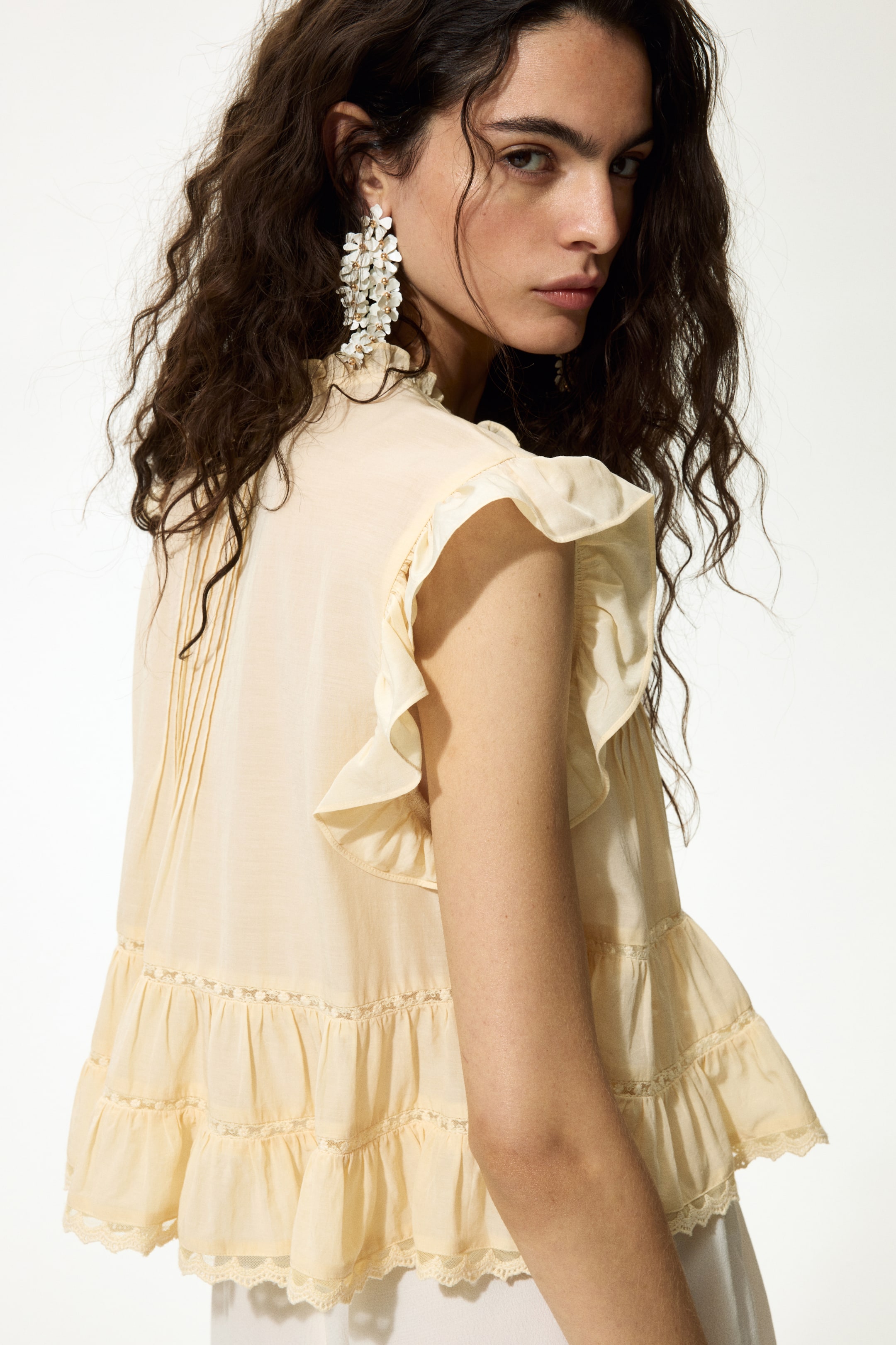 View larger image: The light cream top showcases a ruffled high neck and ruffled cap sleeves. Vertical pin-tucks adorn the upper back, while tiered ruffles, each finished with lace trim, complete the bottom hem.