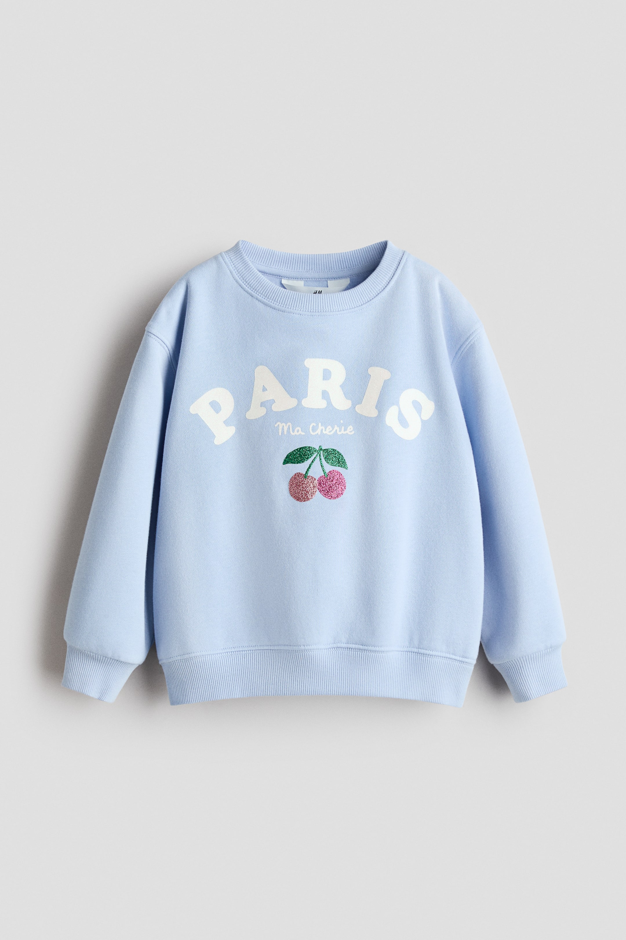 View larger image: A light blue sweatshirt featuring a ribbed crew neck, ribbed cuffs, and a ribbed hem. The front displays "PARIS" in white block letters with "Ma Cherie" in smaller script below, alongside two glittering pink and purple cherries.