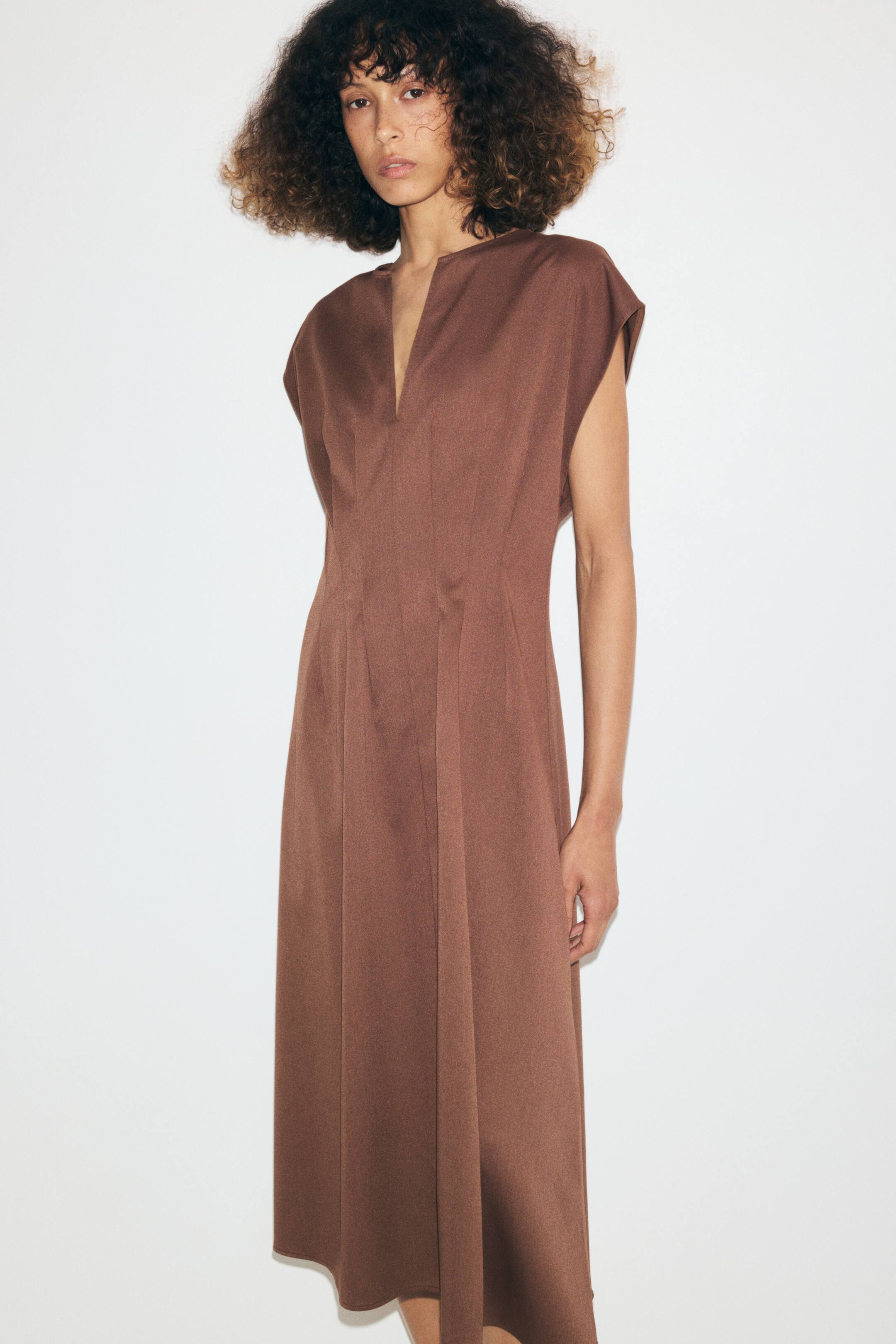 Ampliar la imagen: La modelo lleva puesto un vestido marrón, a la altura de la pantorrilla, confeccionado en twill de caída suave. El vestido presenta un corpiño ajustado con una abertura en forma de V en la parte frontal, mangas tipo capa y pinzas de entalle visibles en la cintura que dan paso a una falda ligeramente acampanada.