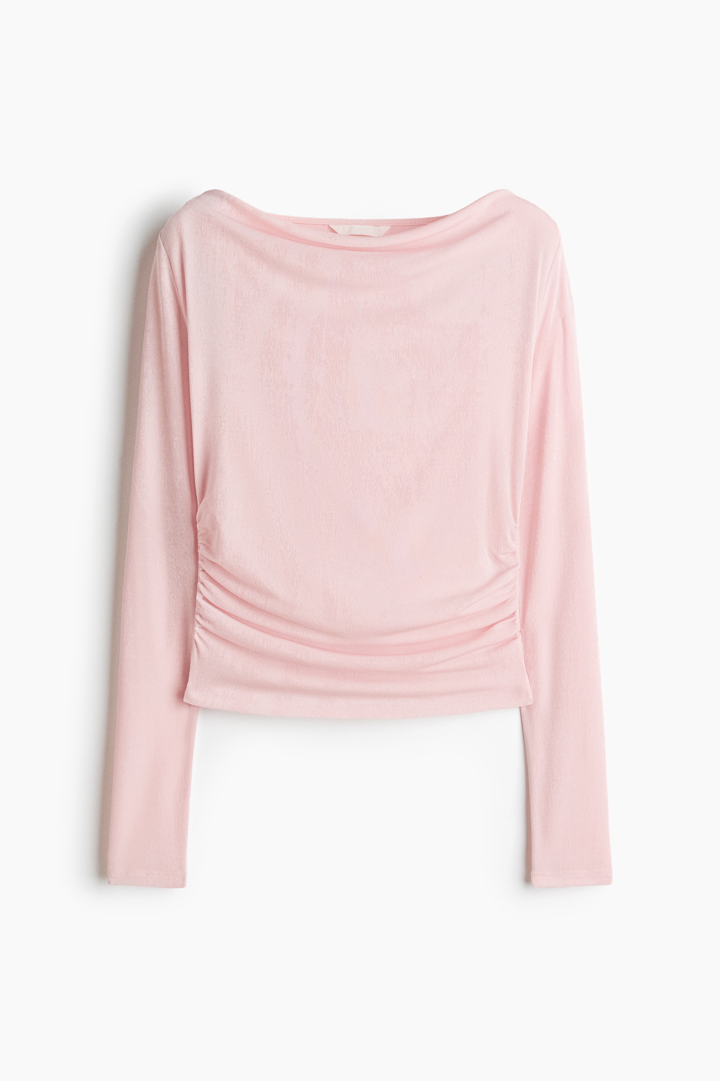 A light pink, long-sleeved top in a fine ribbed jersey with a subtle sheen is worn by the model, featuring a wide scoop neckline and gathered side seams that create a draped effect at the waist. The top is styled with blue denim jeans as the model poses with her hands resting on her hips.