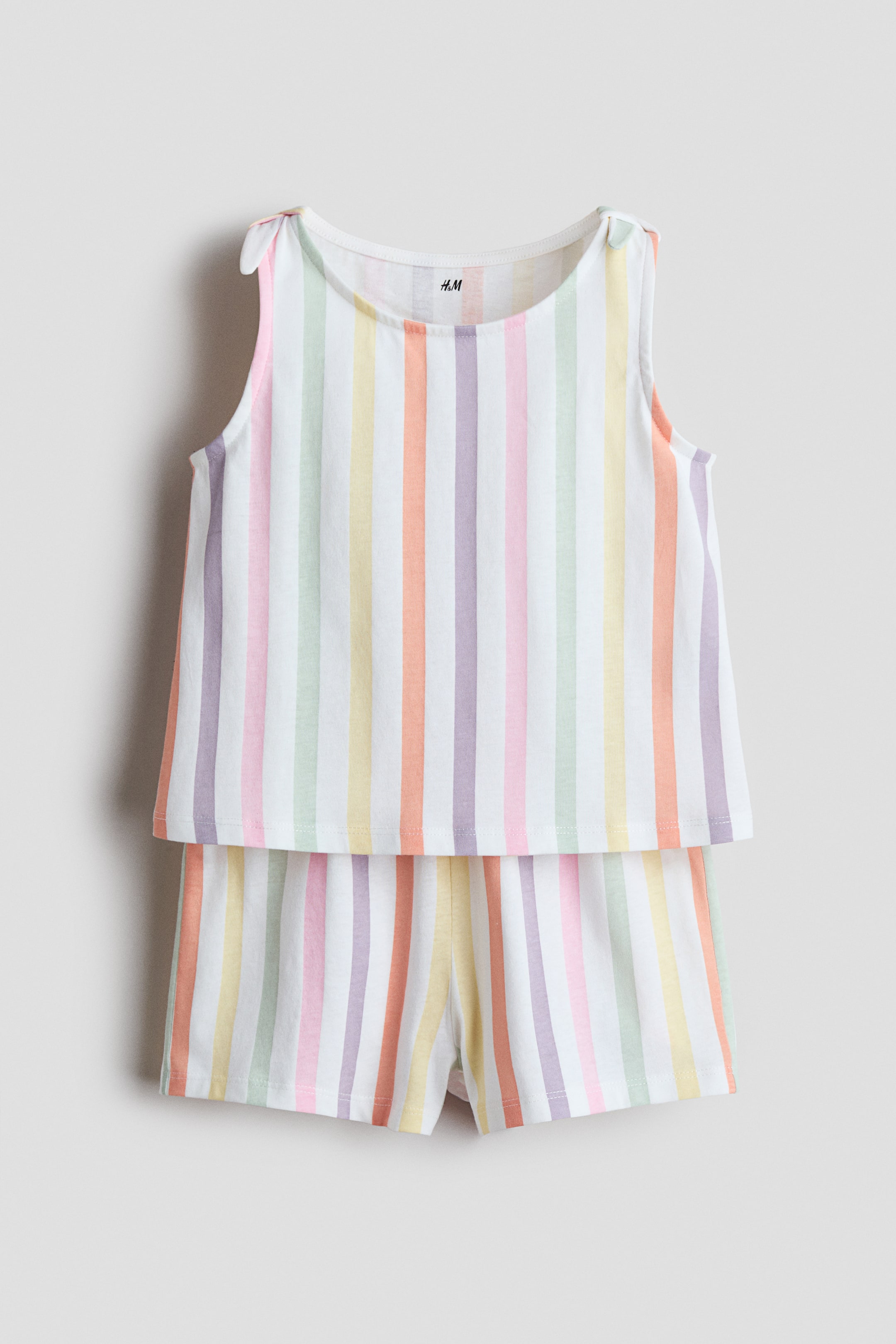 Grotere afbeelding bekijken: Een tweedelig kinderkledingset wordt van voren gepresenteerd, bestaande uit een mouwloze top en bijpassende shorts. De top heeft een ronde hals met strikjes op de schouders, en beide stukken zijn bedrukt met verticale strepen in pastelroze, groen, geel, oranje en paars op een witte achtergrond.