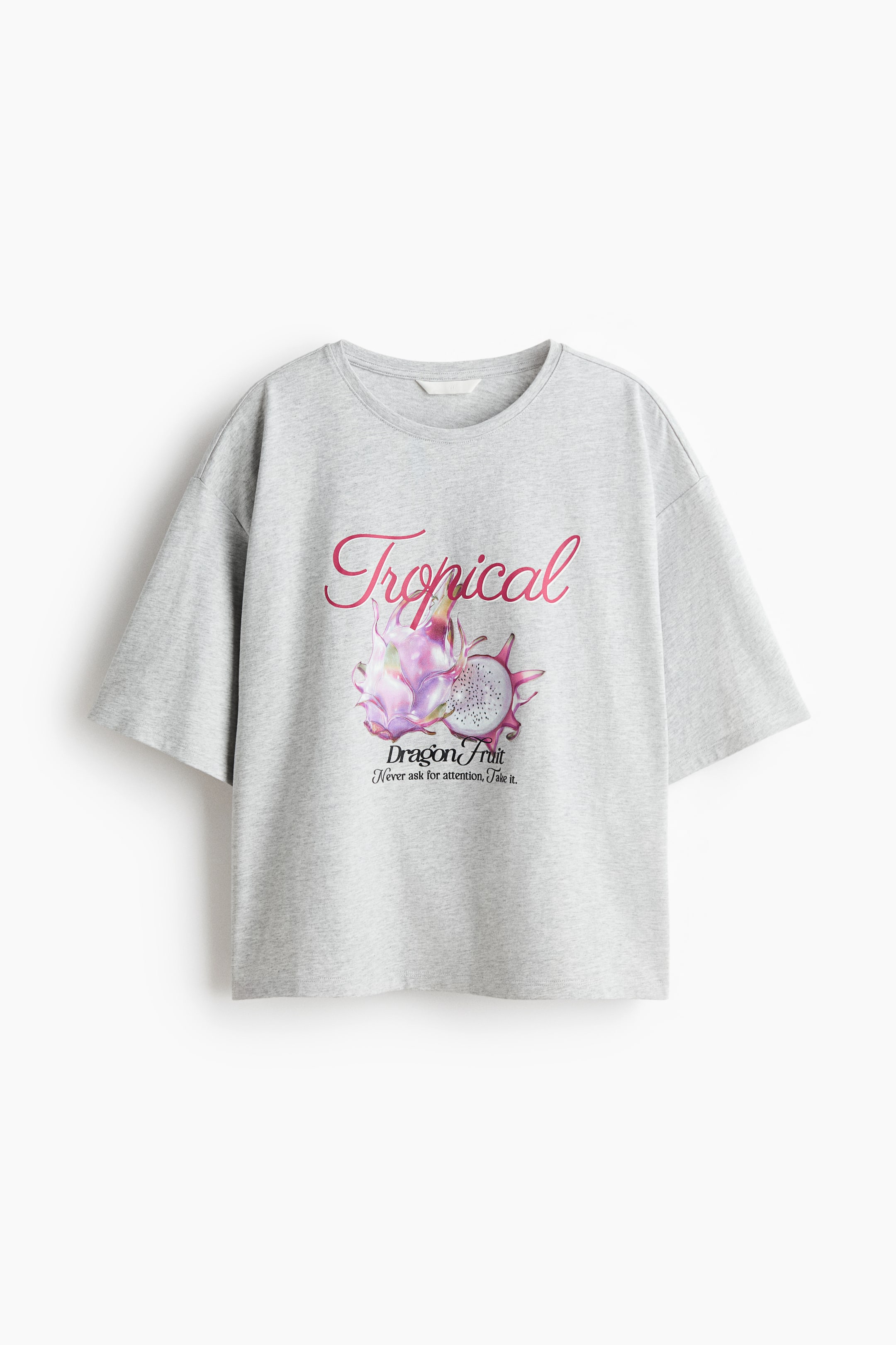 拡大画像を表示: グレーのTシャツは、ラウンドネックラインと短くゆったりとした袖が特徴です。フロントには「Tropical」というピンクのスクリプトで書かれたグラフィックプリントが施されており、その上にはドラゴンフルーツが2つ、1つは丸ごと、もう1つはスライスされて開かれた状態で描かれています。さらに「Dragon Fruit」と「Never ask for attention, take it.」というテキストが添えられています。