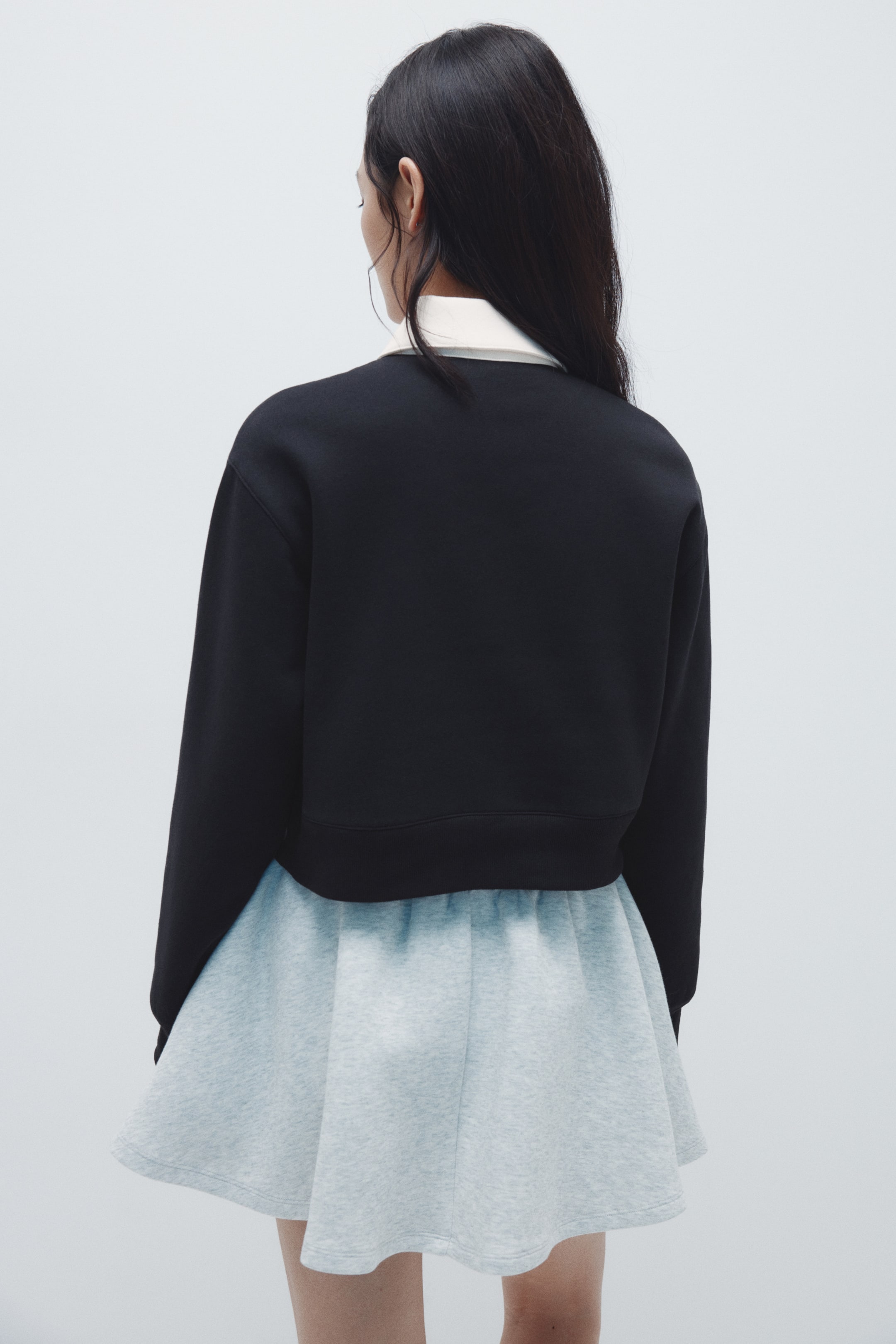 Xem ảnh lớn hơn: Một chiếc áo sweatshirt đen dáng ngắn đang được nhìn từ phía sau, với tay áo dài có cổ tay và gấu áo được may viền. Một chiếc cổ áo trắng được thấy rõ bên dưới cổ áo tròn của nó.