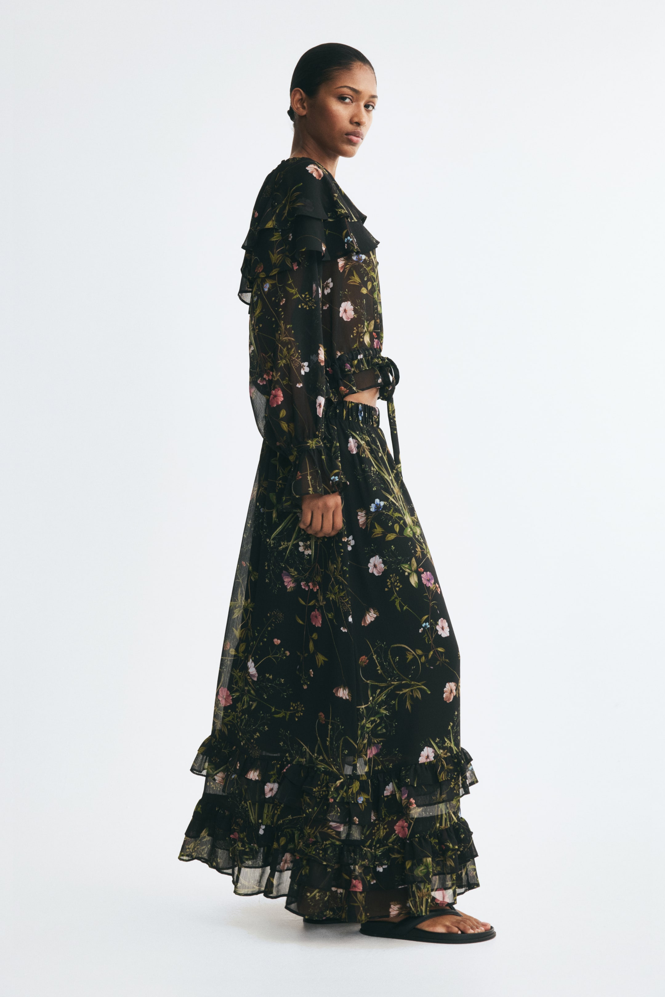 View larger image: A calf-length, slightly A-line skirt in black crinkled chiffon with a floral pattern is being worn by the model. The skirt features an elasticated waist and frill-trimmed tiers above the hem, and is paired with a matching long-sleeve floral blouse.