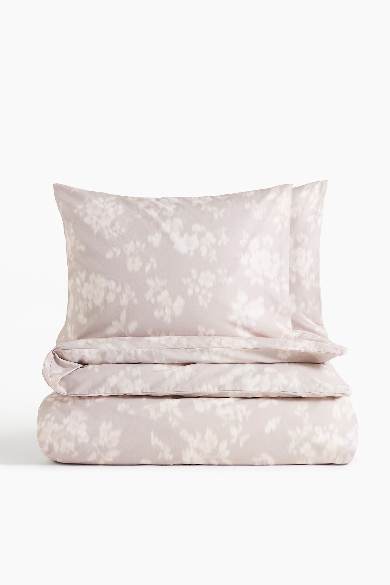 Ein Bettbezug-Set aus Baumwoll-Perkal in einem dezenten Rosé-Beige zeigt aus frontaler Perspektive einen sanft verschwommenen weißen Blumenprint. Das Set umfasst zwei passende Kissenbezüge, die aufrecht hinter einem ordentlich gefalteten Bettbezug stehen und die glatte Textur des Stoffes hervorheben.