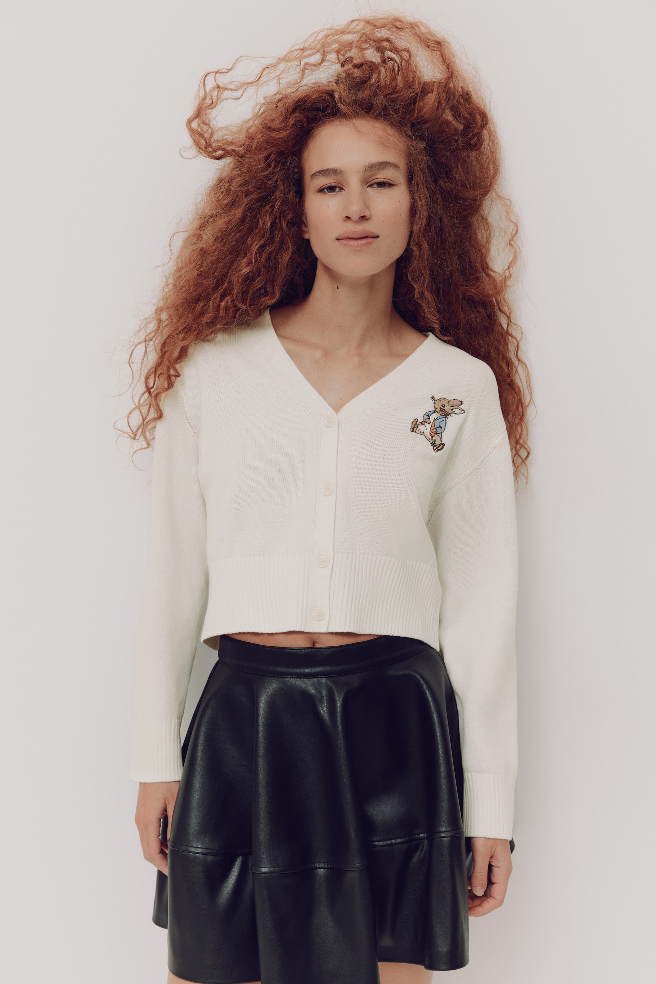 이미지 확대 보기: 화이트 크롭 카디건으로 V넥과 연한 색 버튼 5개가 있는 앞면 버튼 여밈이 특징입니다. 골지 소매단과 골지 밑단이 적용된 긴 소매를 갖추고 있으며, 왼쪽 가슴에 자수 처리된 Peter Rabbit 모티프가 들어가 있습니다.