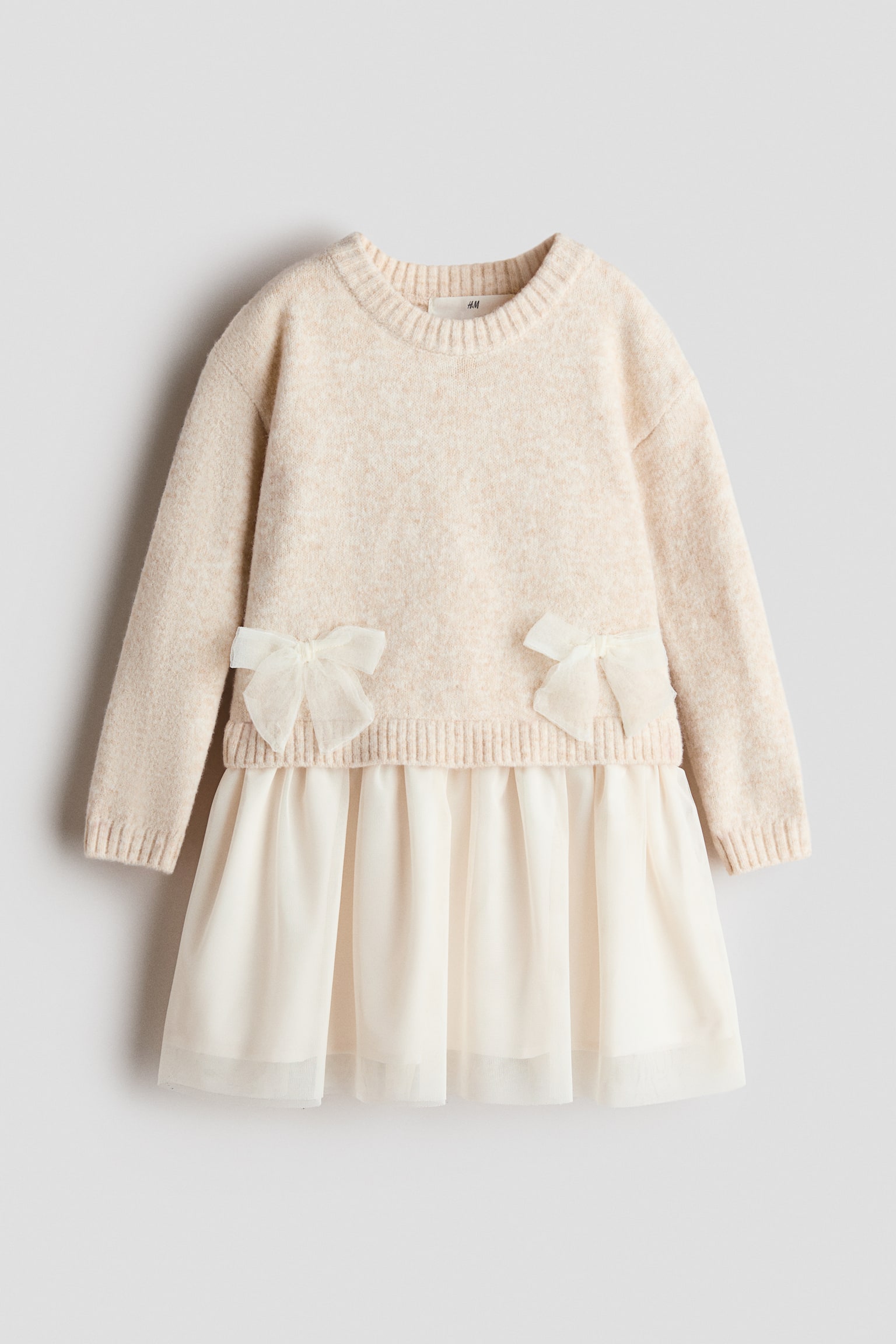 A long-sleeved knit dress is shown facing front, featuring a light beige sweater-style bodice with a ribbed crew neck, ribbed cuffs, and two decorative cream tulle bows at the waist. The bodice transitions into a flared, layered off-white tulle skirt.