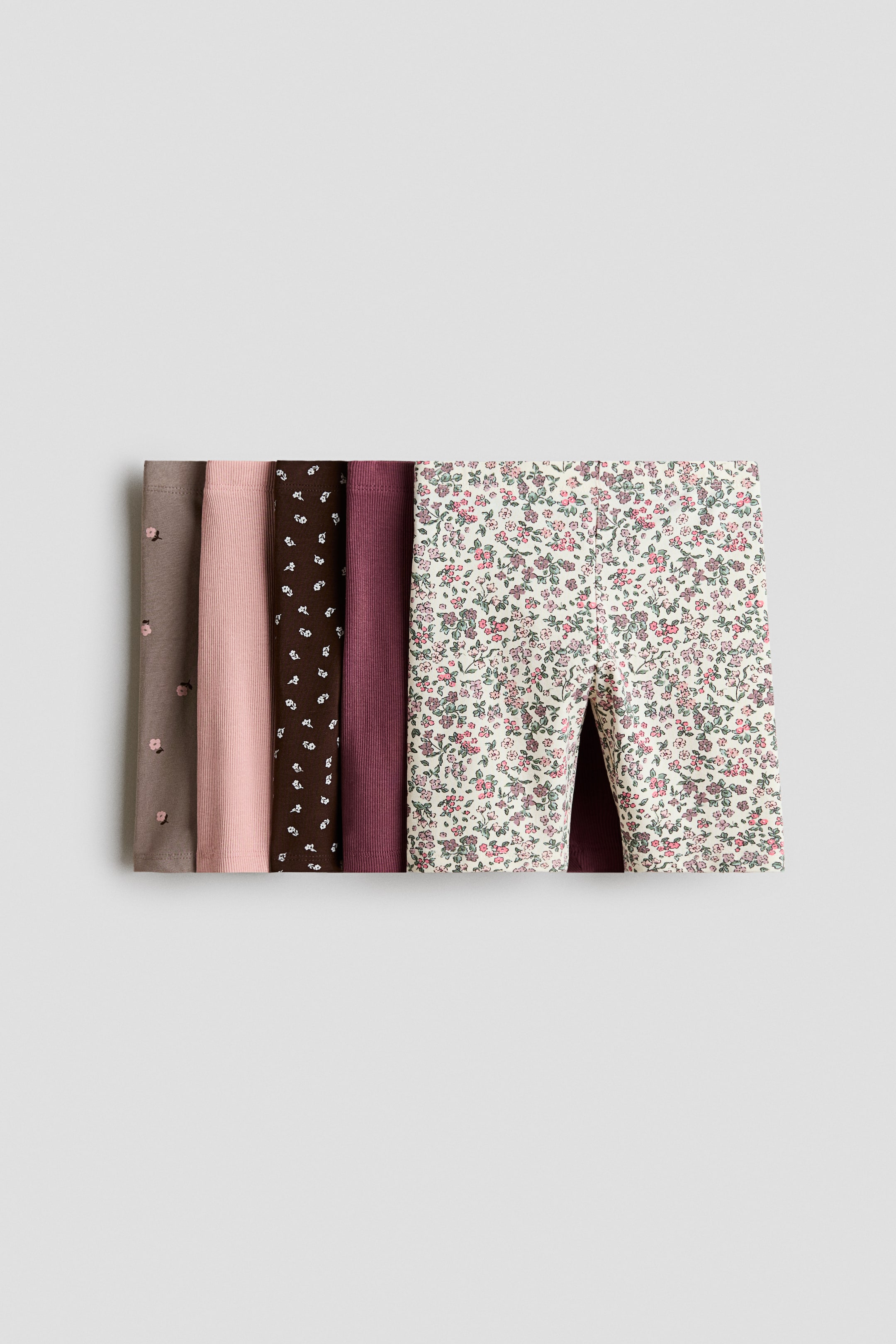 View larger image: A five-pack of children’s bike shorts is laid flat, front-facing, showcasing a variety of colours and patterns. The top pair features an all-over pink and green floral pattern, while the layered pairs beneath feature a solid ribbed plum, a dark brown floral print, a solid ribbed light pink, and a taupe fabric with small pink floral buds.