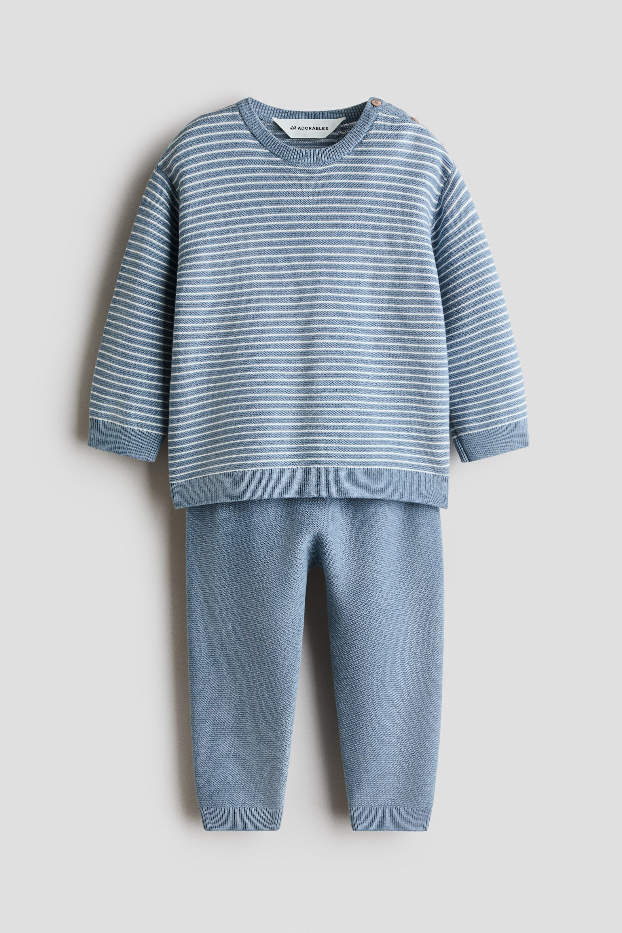 View larger image: A knitted baby outfit featuring a light blue long-sleeved top with thin white horizontal stripes, ribbed cuffs, and a ribbed hem, along with two small brown buttons on the left shoulder. Matching solid light blue knitted trousers complete the set, showcasing ribbed ankles.