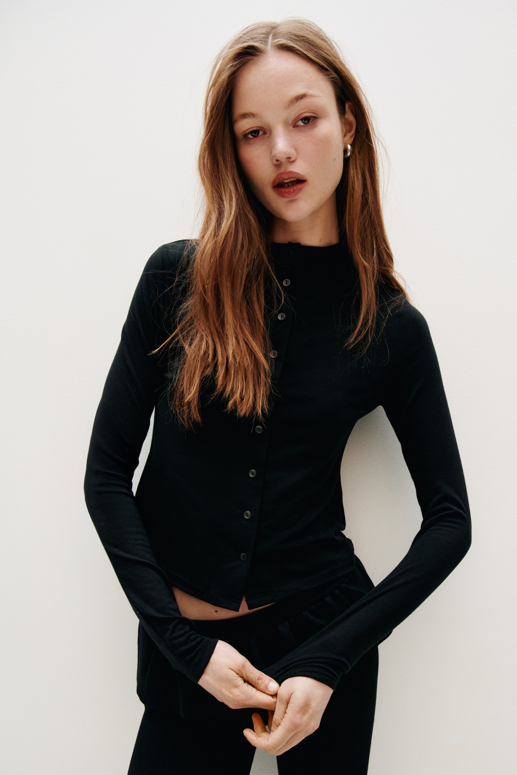 Grösseres Bild ansehen: Ein schwarzer, figurbetonter Jersey-Cardigan mit kleinem Stehkragen und Knöpfen vorne wird vom Model getragen. Der Cardigan zeigt lange Ärmel und ist unten teilweise aufgeknöpft, wodurch ein leichter Bauchansatz sichtbar wird.