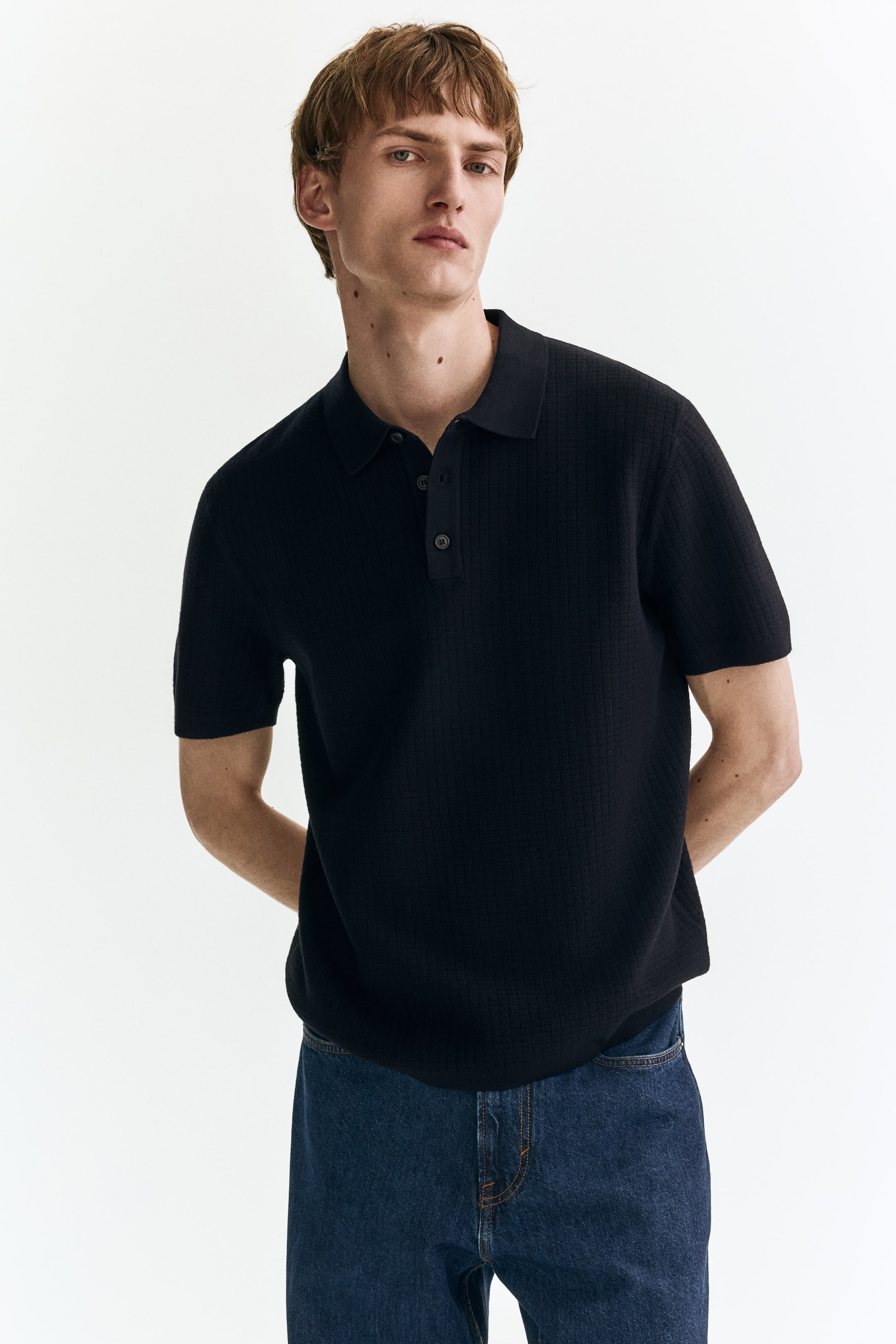View larger image: A black knitted polo shirt is showcased, featuring a subtle textured grid pattern throughout its fabric. It is crafted with a pointed collar, a three-button placket, and short sleeves, presenting both a front and side view.