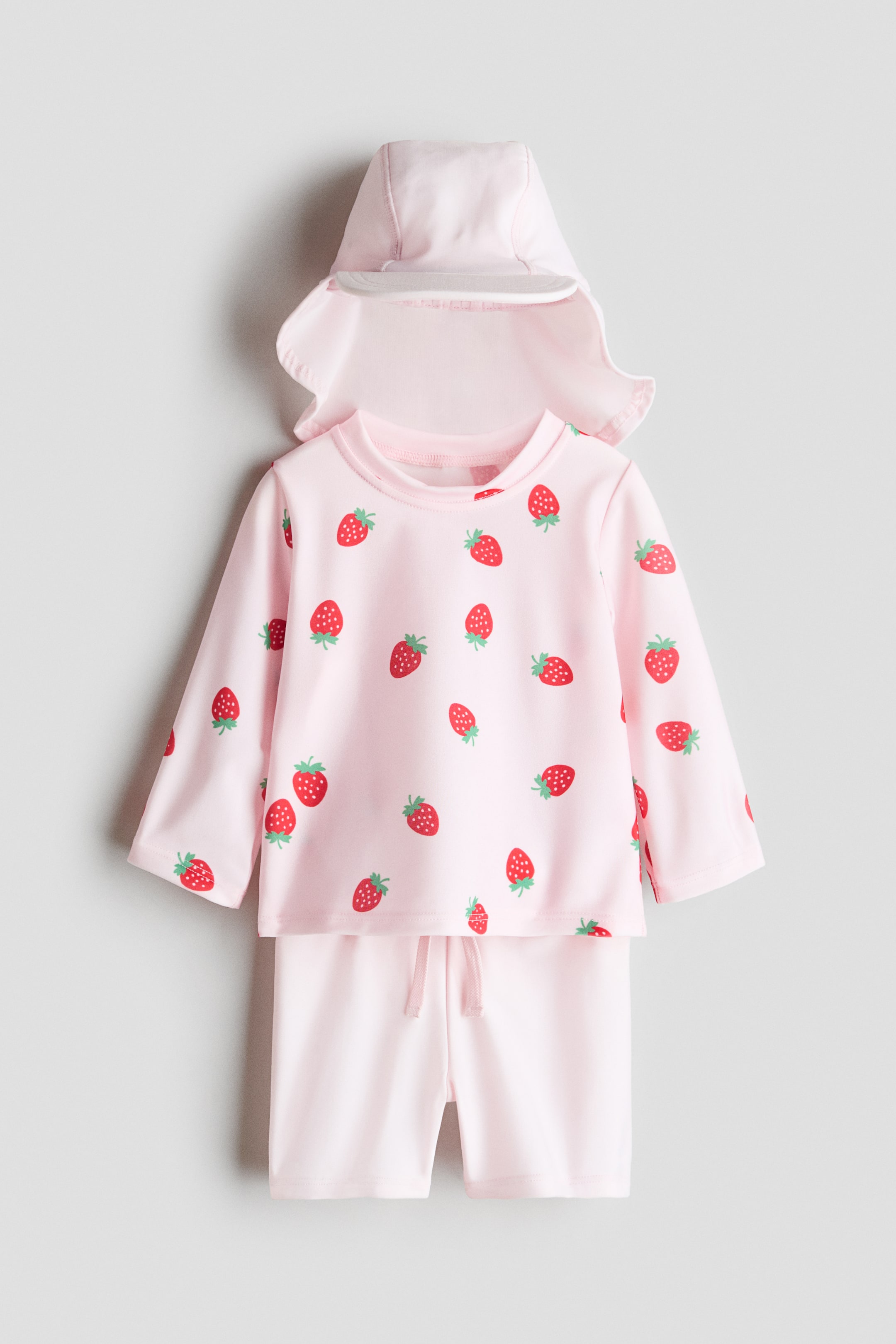 Zobacz większy obraz: Zestaw kąpielowy w jasnoróżowym kolorze z ochroną UV jest prezentowany z przodu. Składa się z bluzki z długimi rękawami, okrągłym dekoltem, rękawami raglanowymi i czerwonym wzorem truskawek, a także z solidnych różowych szortów z elastycznym paskiem i sznurkiem w pasie oraz pasującej czapki z daszkiem z przodu i klapką z tyłu.