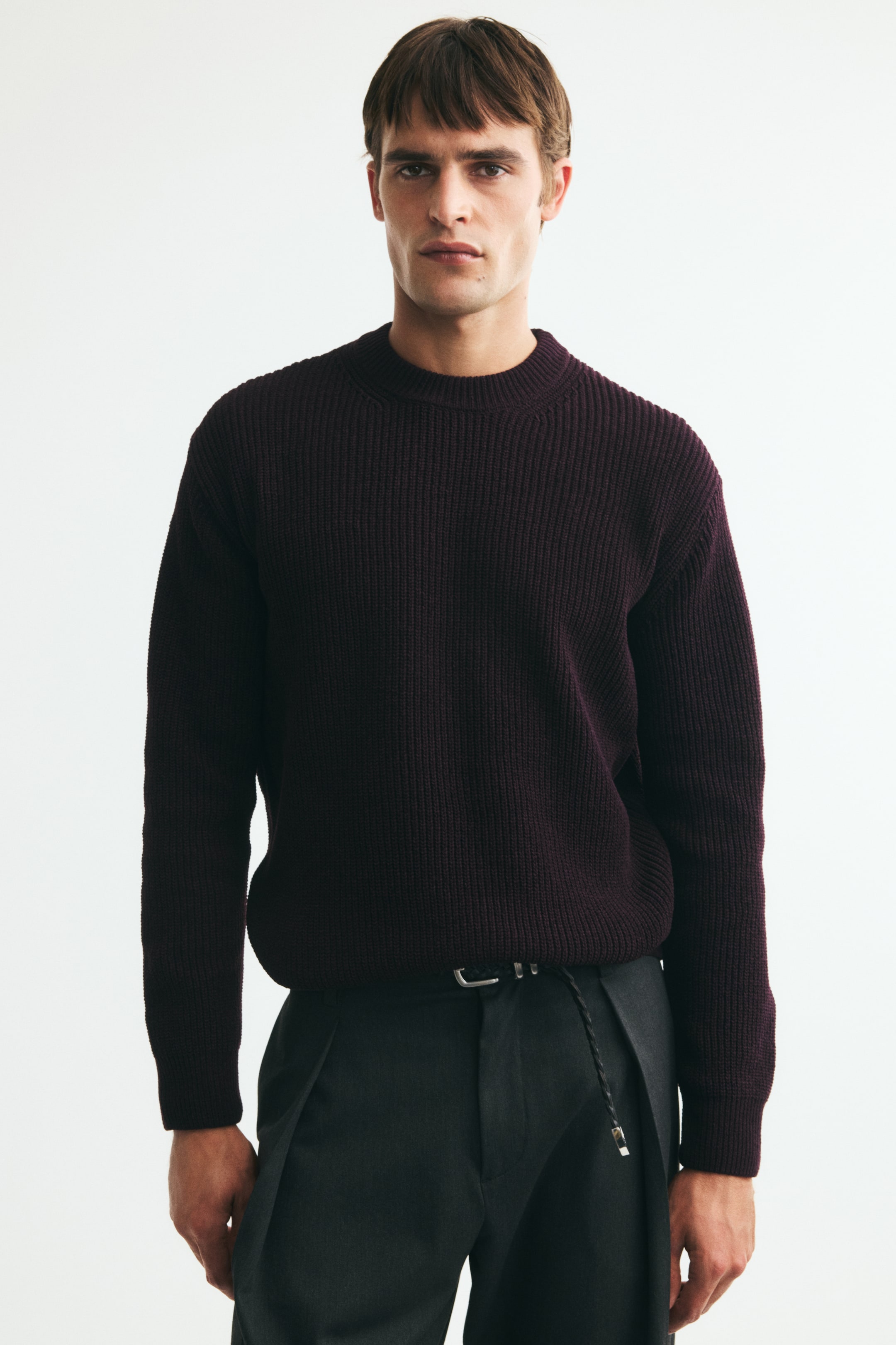 View larger image: The model is wearing a dark burgundy rib-knit sweater featuring a relaxed fit, crew neckline, dropped shoulders, and long sleeves. The sweater is styled with a slight tuck into dark trousers at the waist.