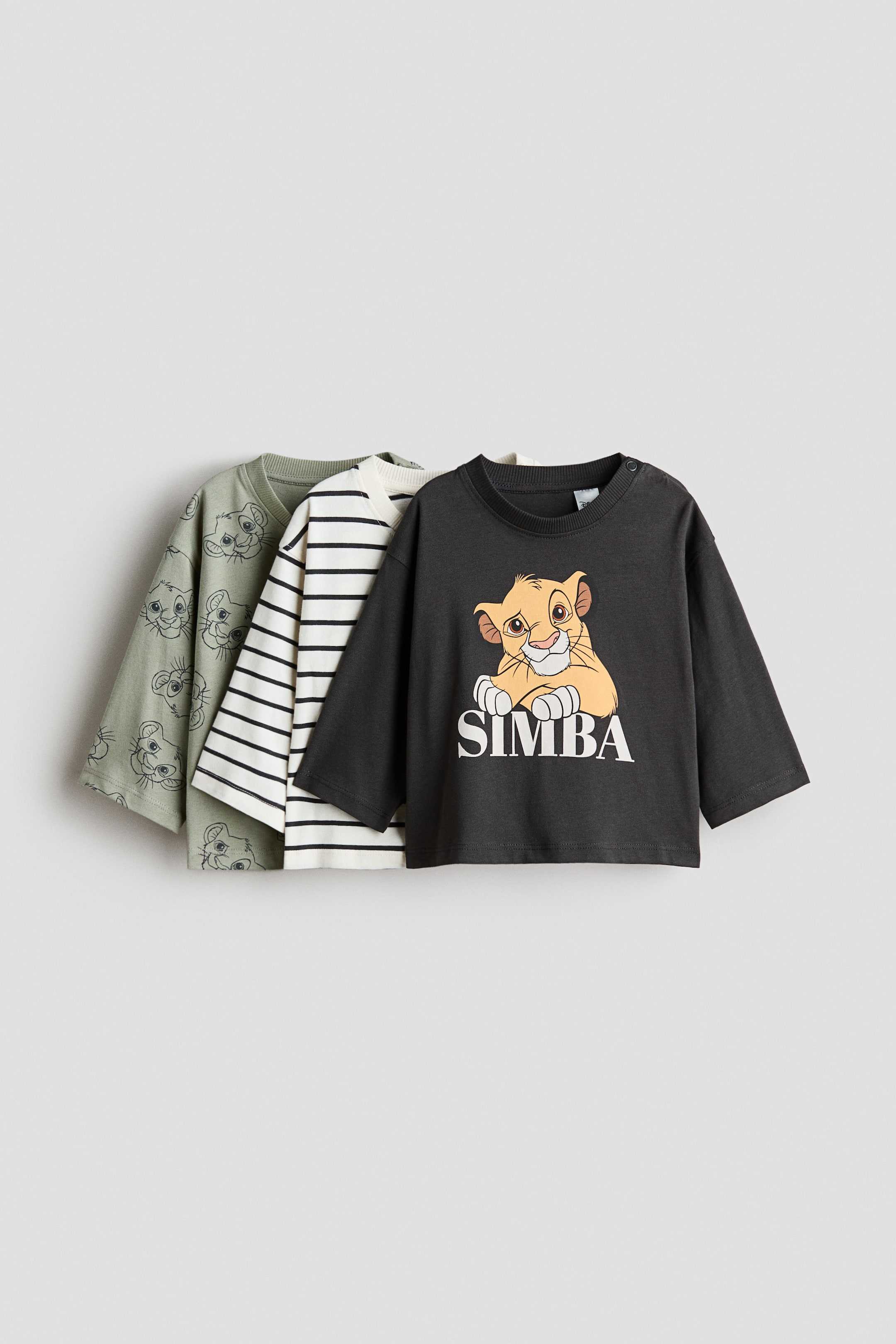 View larger image: A set of three long-sleeved T-shirts is being presented, layered from right to left, all facing forward. The foremost dark grey T-shirt features a young lion graphic with “SIMBA” text and a crew neck with a snap-button closure. Behind it, a cream T-shirt with black horizontal stripes and a sage green T-shirt with an all-over lion-face outline print are visible.