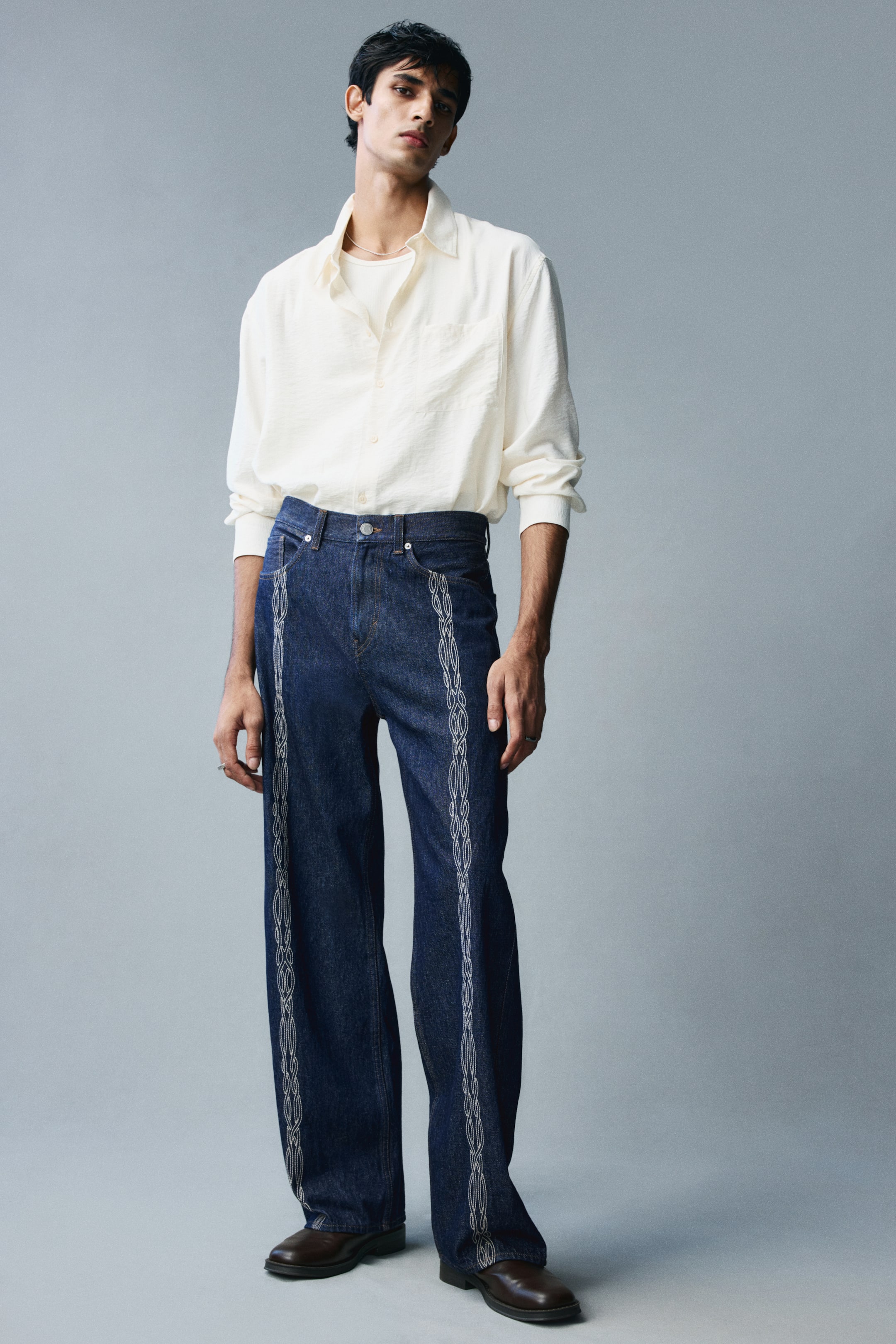 View larger image: Dark blue, loose-fit denim jeans featuring a straight-leg silhouette and vertical chain-stitch embroidery running down the front of each leg are being worn by the model. The regular-waisted jeans are styled with a cream-coloured button-up shirt tucked into the waistband.