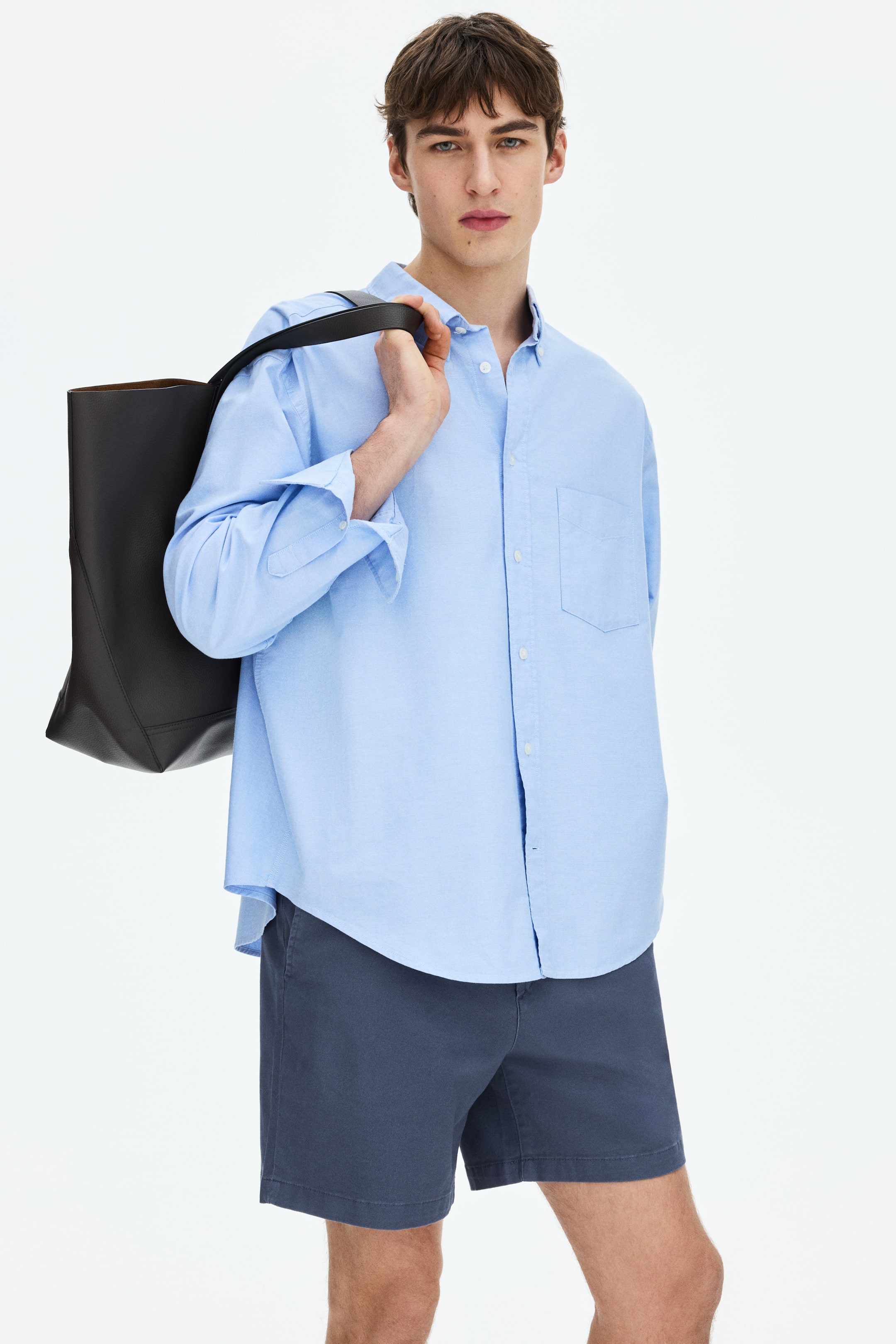 View larger image: The light blue button-down shirt features a button-down collar, a full button placket with white buttons, and a single chest pocket on the left side. Its long sleeves are rolled up to the elbows, and it is worn untucked over dark shorts, showcasing a curved hem.
