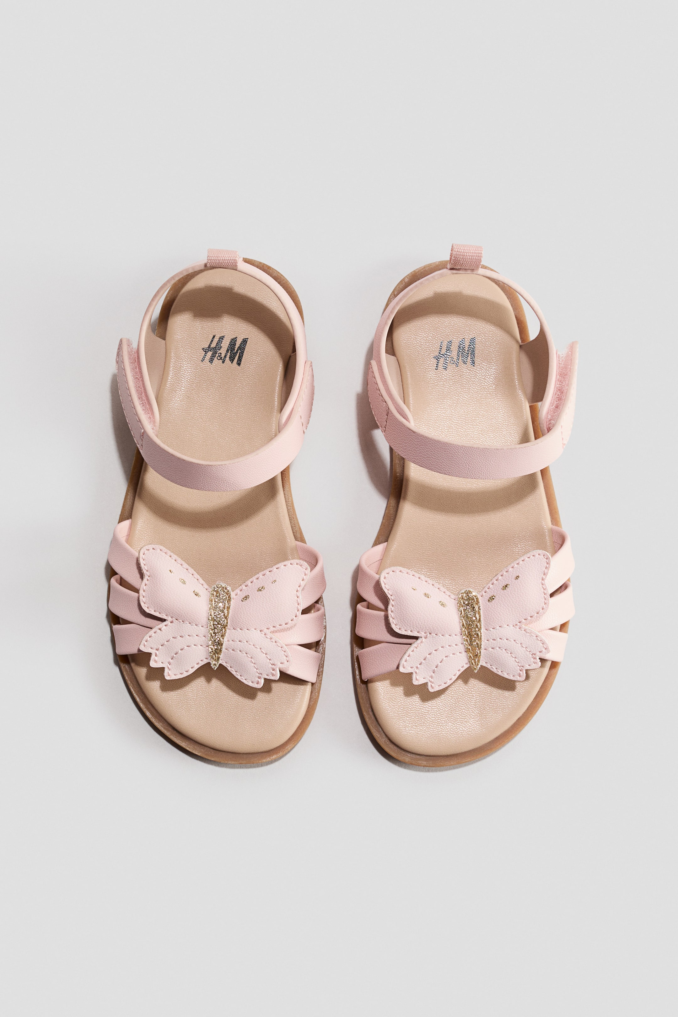 View larger image: Two pink children's sandals are showcased from a top-down perspective, each featuring multiple toe straps, an adjustable ankle strap with a Velcro fastening, and a butterfly appliqué with gold glitter detailing on the front. The H&M logo is visible on the insole of each sandal.