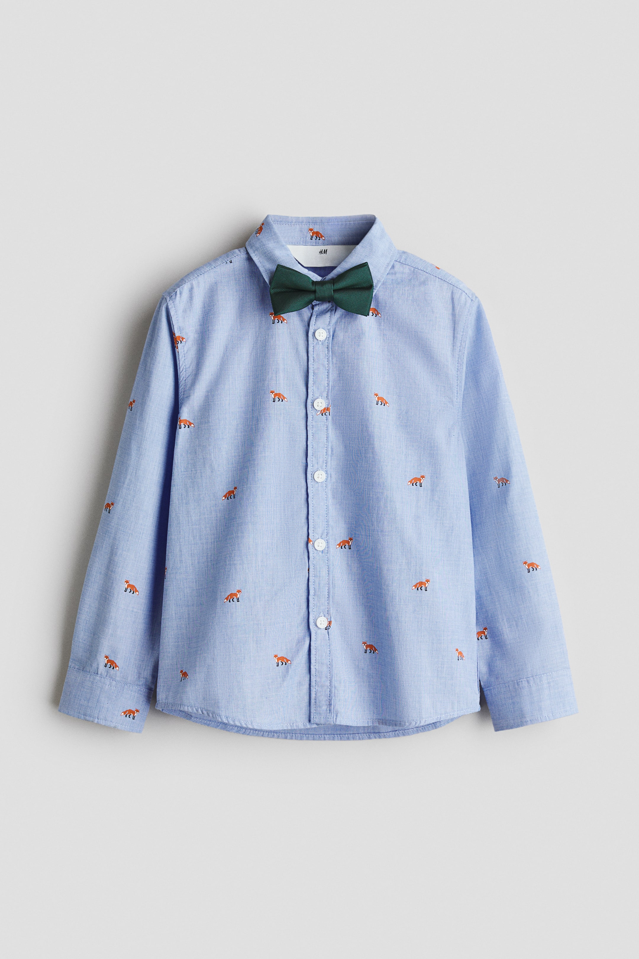 View larger image: A light blue long-sleeved shirt showcases a repeating pattern of small orange fox motifs, featuring a classic pointed collar with an attached dark green bow tie and white buttons fastening down the front placket.