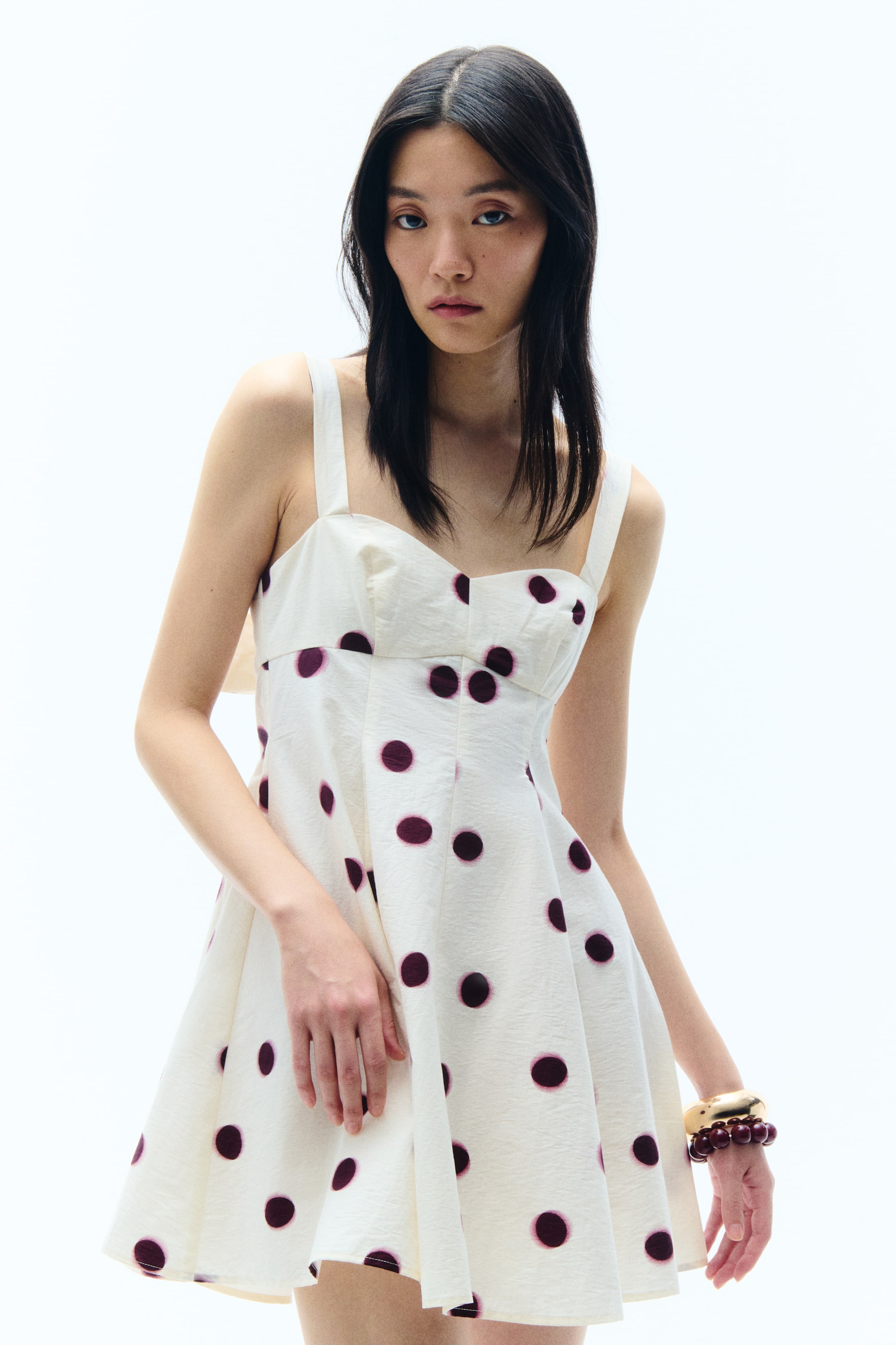 View larger image: An off-white dress featuring a sweetheart neckline with thick shoulder straps and a fitted waist. The silhouette flows into a flared skirt, detailed with large, irregularly spaced purple polka dots and a subtle crinkled fabric texture.
