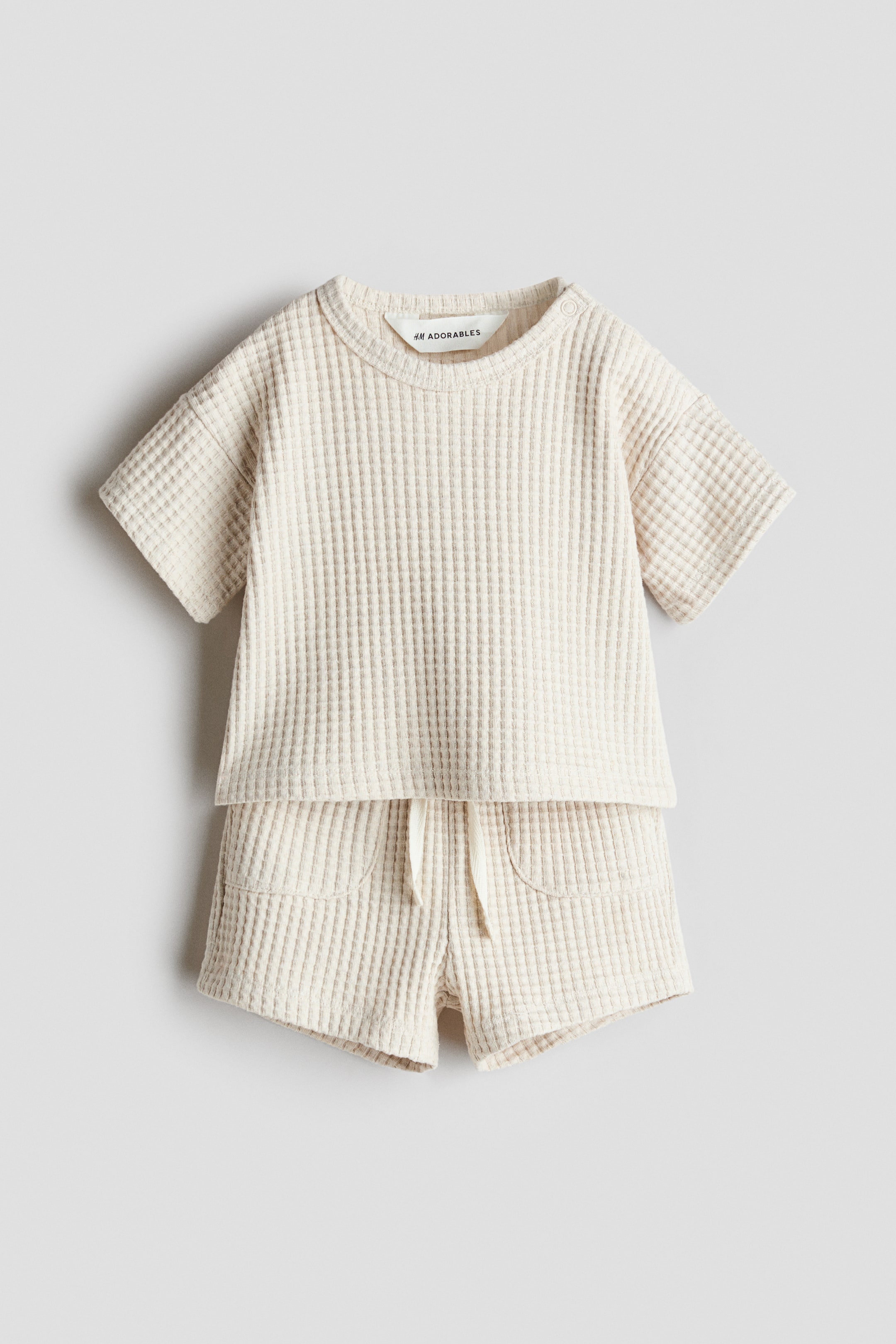 View larger image: An off-white, waffled jersey set is featuring a top and shorts, laid flat as a front view. The top is designed with a round, trimmed neckline, a press-stud on one side, dropped shoulders, and short sleeves, and the matching shorts are showing an elasticated drawstring waist with a visible drawstring and two patch front pockets.
