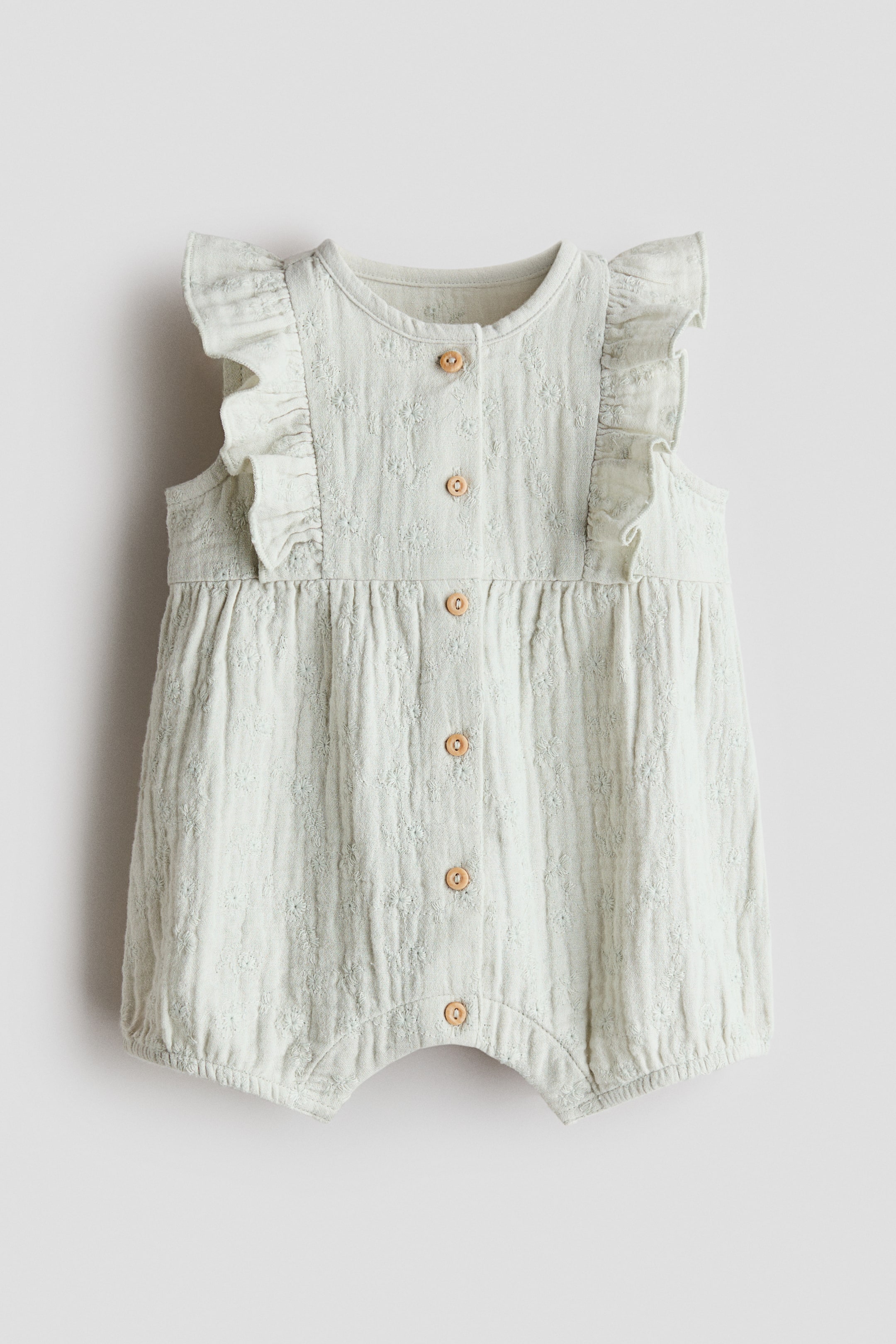 View larger image: This sleeveless romper suit showcases its front view, crafted from light green cotton muslin with broderie anglaise detailing. It features a round neckline, wooden buttons running down the front, flounces over the shoulders, and gentle elastic at the leg openings.