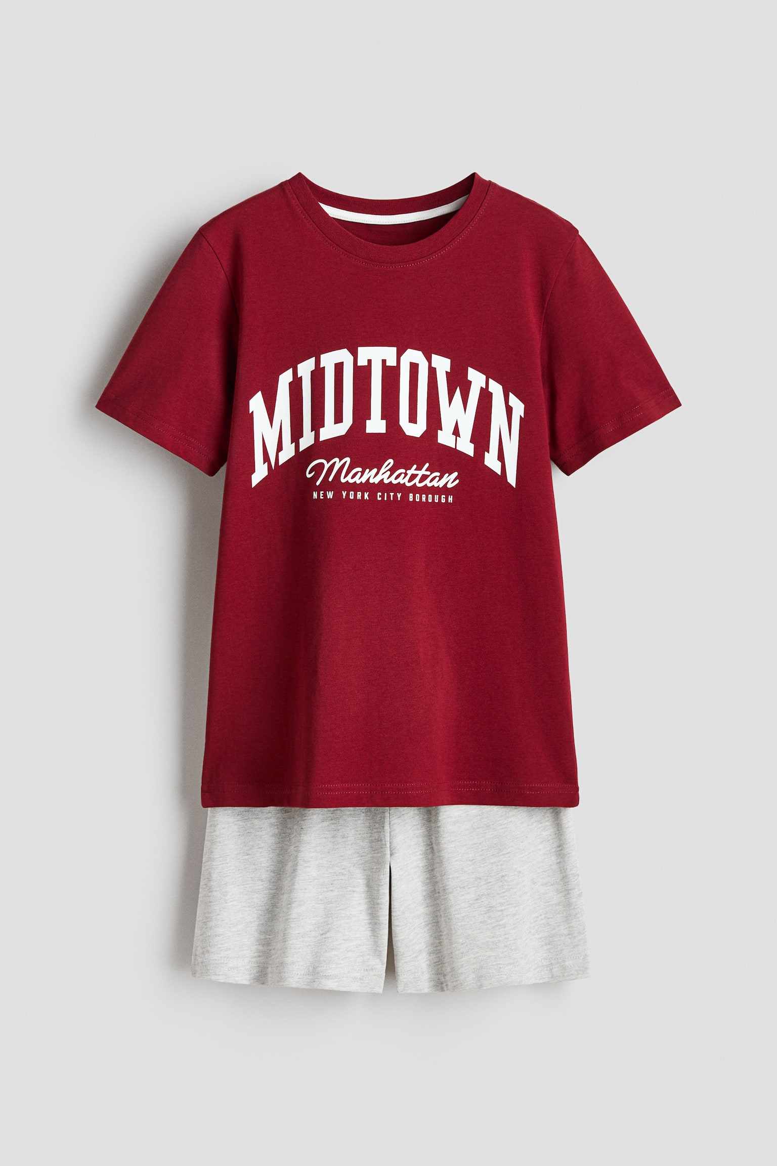 A pyjama set is shown from the front, highlighting a maroon tee with a round, ribbed neckline, subtly dropped shoulders, and a white 'MIDTOWN Manhattan NEW YORK CITY BOROUGH' print. Below, light grey shorts are seen, featuring an elastic waistband.
