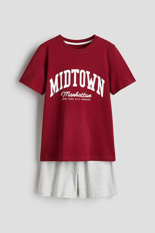 Ein Pyjama-Set zeigt seine Vorderansicht, bestehend aus einem bordeauxroten T-Shirt mit einem runden, gerippten Halsausschnitt, sanft fallenden Schultern und einem weißen Aufdruck "MIDTOWN Manhattan NEW YORK CITY BOROUGH". Darunter sind hellgraue Shorts zu sehen, die über einen elastischen Bund verfügen. - 1