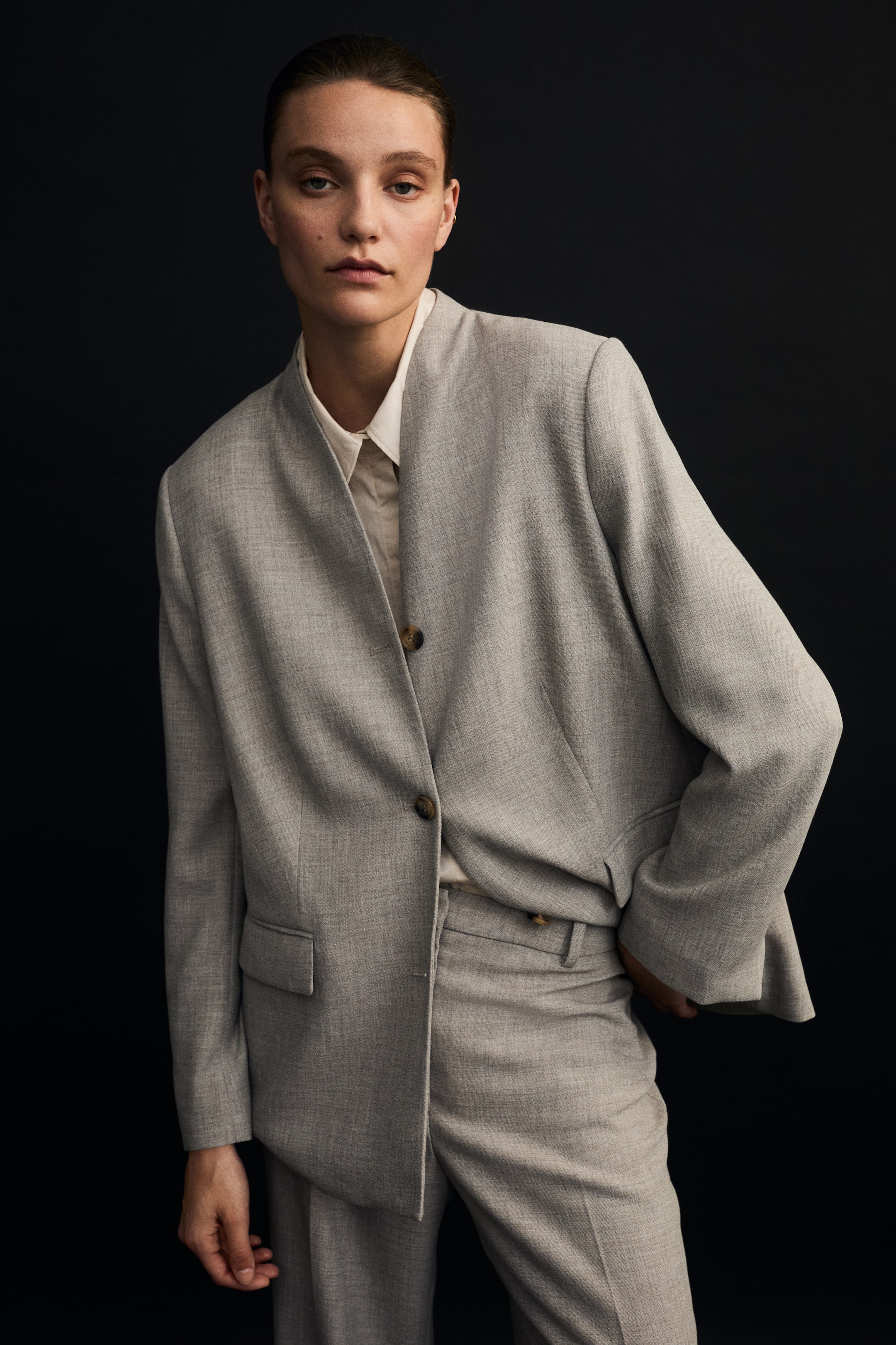 View larger image: A light grey blazer and matching trousers are showcased from a gentle front-left angle, emphasising their laid-back fit and structure. The blazer features a band collar, two brown buttons, and flap pockets, and is worn over an off-white collared shirt, while the matching trousers appear in a relaxed, straight-leg style.
