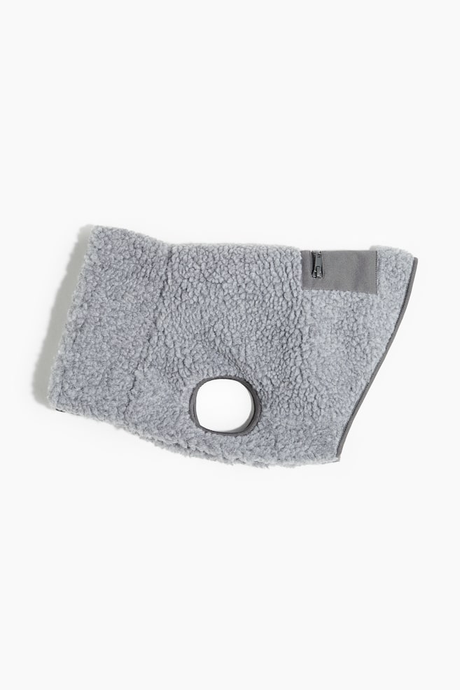 Une veste pour chien grise, faite de tissu en peluche douce, est présentée. Elle présente des détails en tissu tissé gris foncé, y compris un panneau avec une fermeture éclair le long de son côté droit, une ouverture circulaire près du haut pour une laisse, et une plus grande ouverture ovale vers le bas à gauche pour une patte avant. - 1