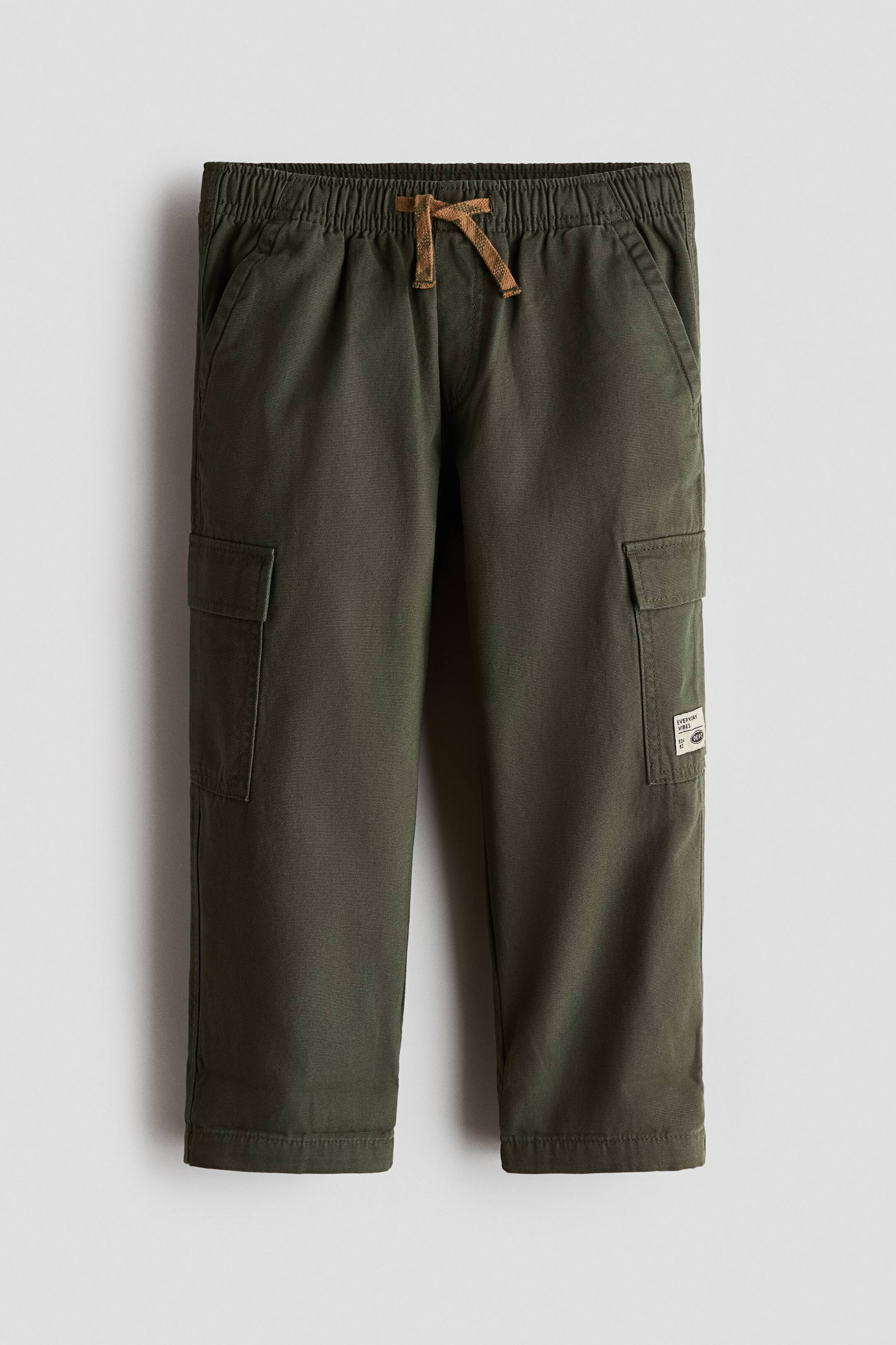 Xem ảnh lớn hơn: Quần jogger cargo màu xanh ô liu bằng vải canvas cotton mềm được chụp từ phía trước, với phom dáng rộng, cạp chun có dây rút màu nâu, túi bên chéo, đường khóa giả và túi patch có nắp trên cả hai ống quần.