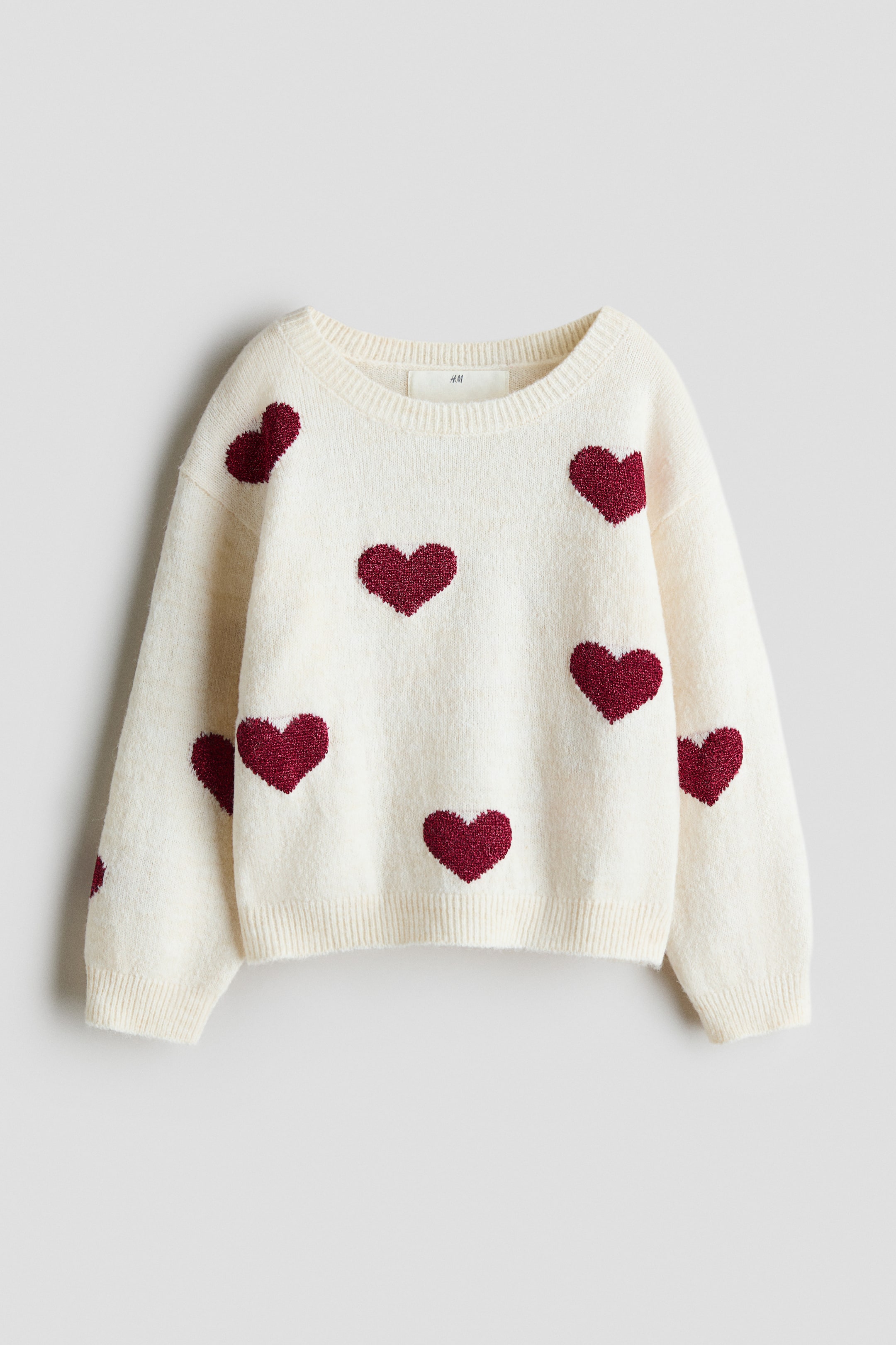 View larger image: A cream-coloured knit sweater is shown from the front, featuring scattered, textured red heart patterns across the body and sleeves. The garment displays a round, rib-trimmed neckline, dropped shoulders, and ribbing at the cuffs and hem.