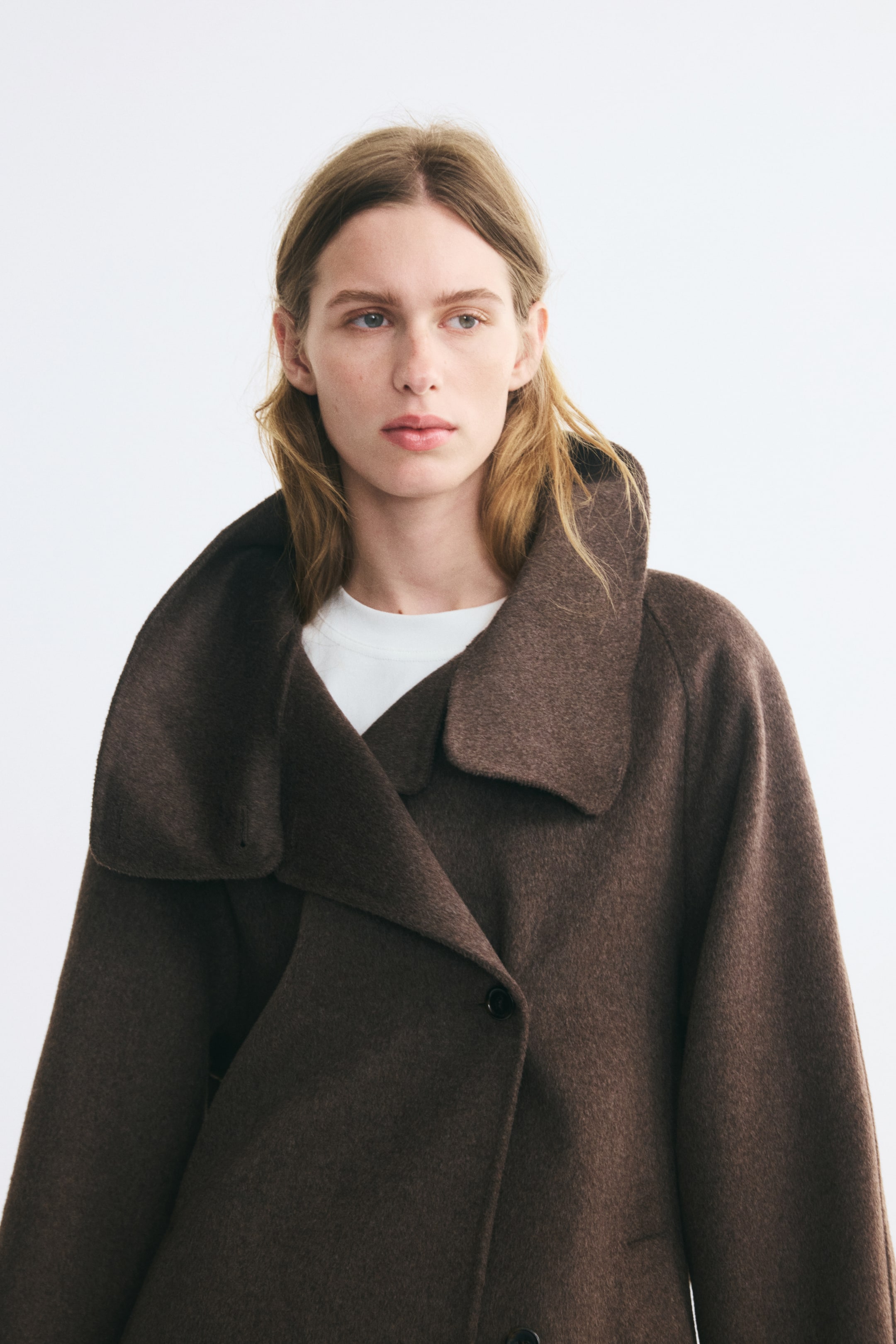 Grotere afbeelding bekijken: Een oversized bruine jas van een gevilte wolmix wordt door het model gedragen over een wit T-shirt. De jas heeft een brede, asymmetrische kraag met trechtervorm, raglanmouwen en een asymmetrische knoopsluiting aan de voorkant.