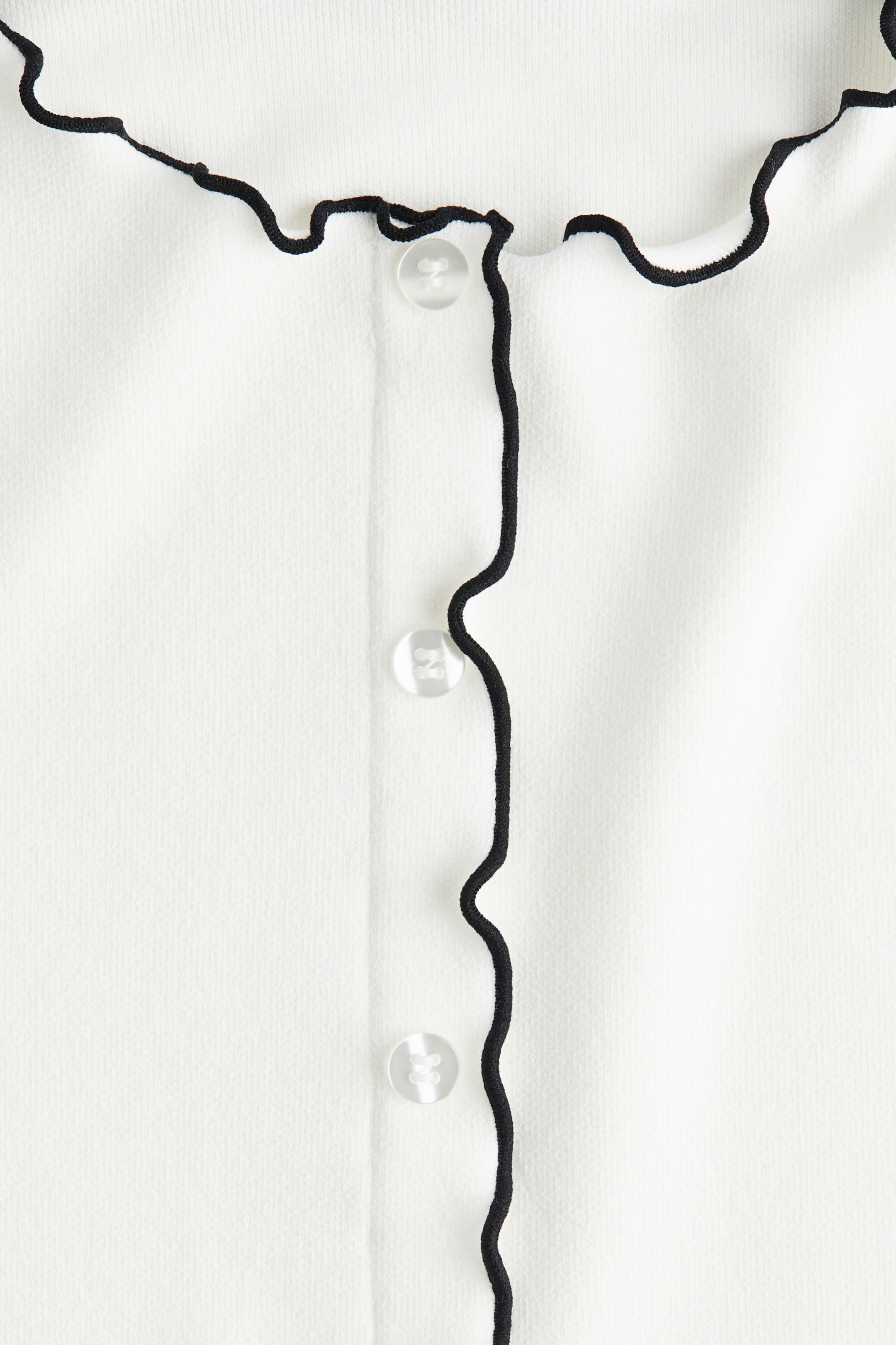 View larger image: A white garment is displaying a black wavy trim along its front placket and neckline, with clear round buttons fastening the front closure.