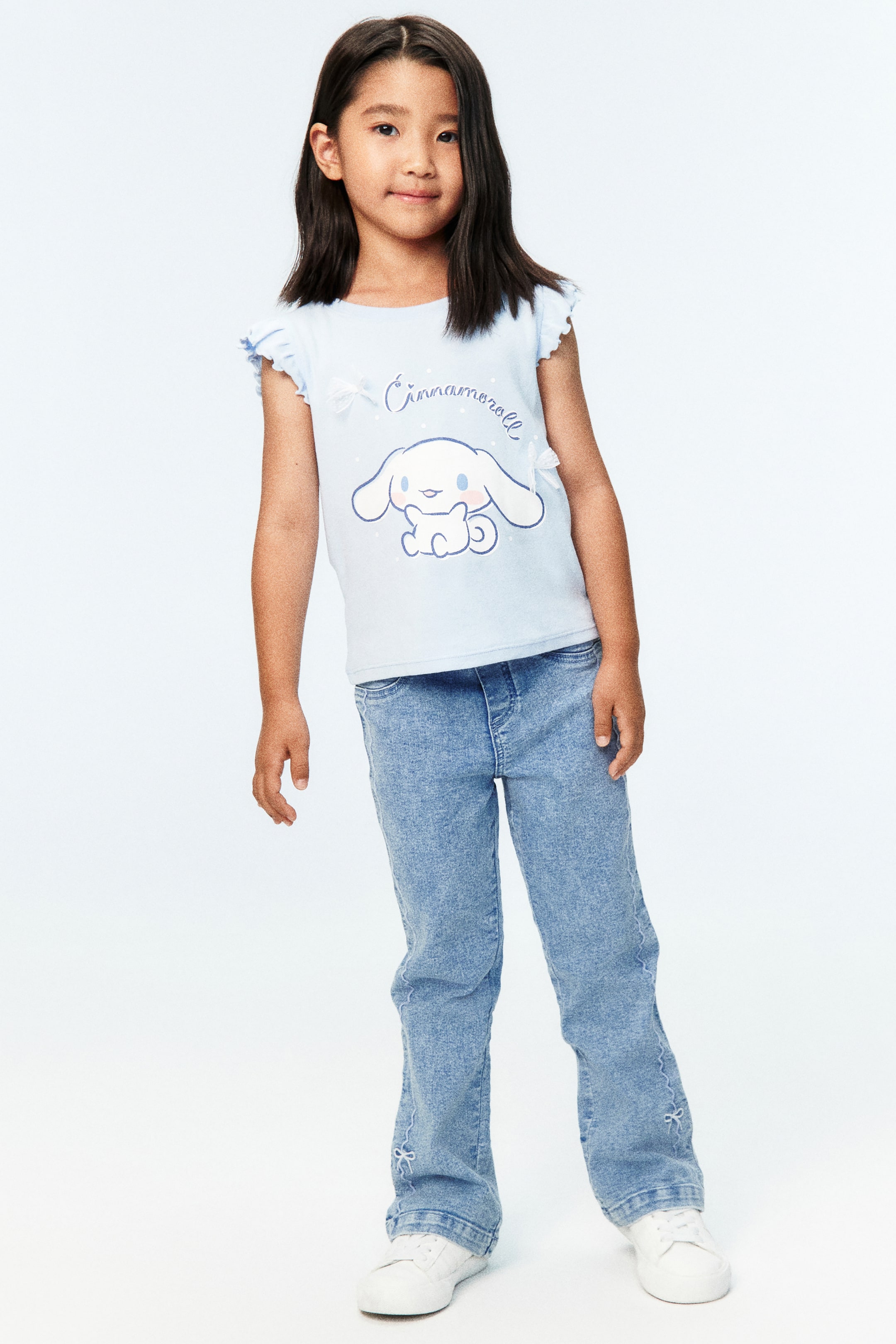 View larger image: The model is wearing light blue denim jeans featuring an elasticated waistband, a regular fit through the hip and thigh, and flared legs with embroidered bow details near the ankles.