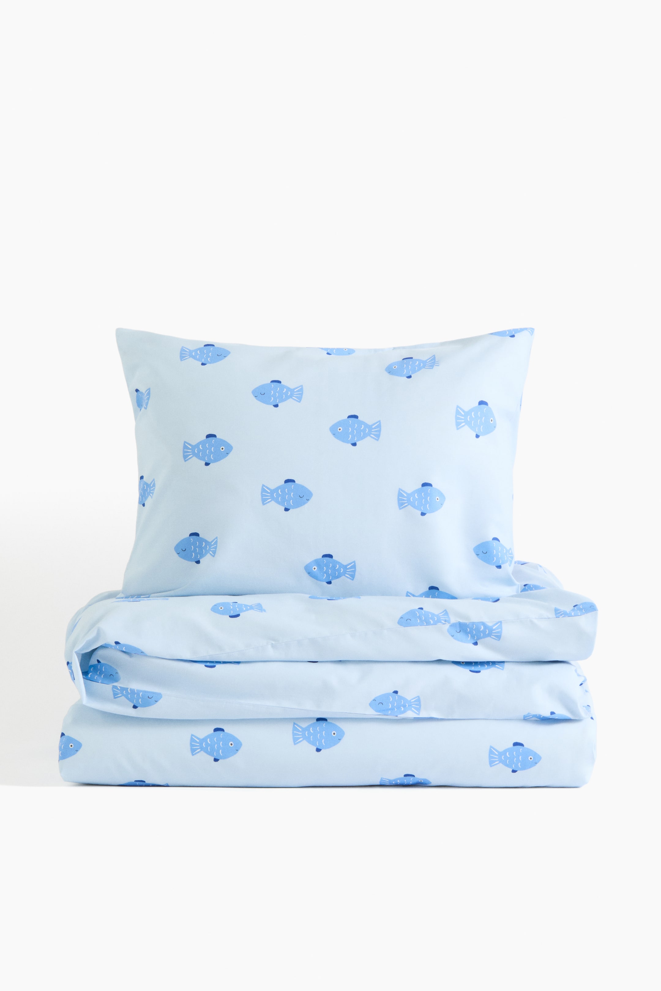 View larger image: A light blue bedding set showcases a pillow standing upright and a duvet cover folded horizontally beneath it, both adorned with an all-over pattern of light and dark blue cartoon fish.