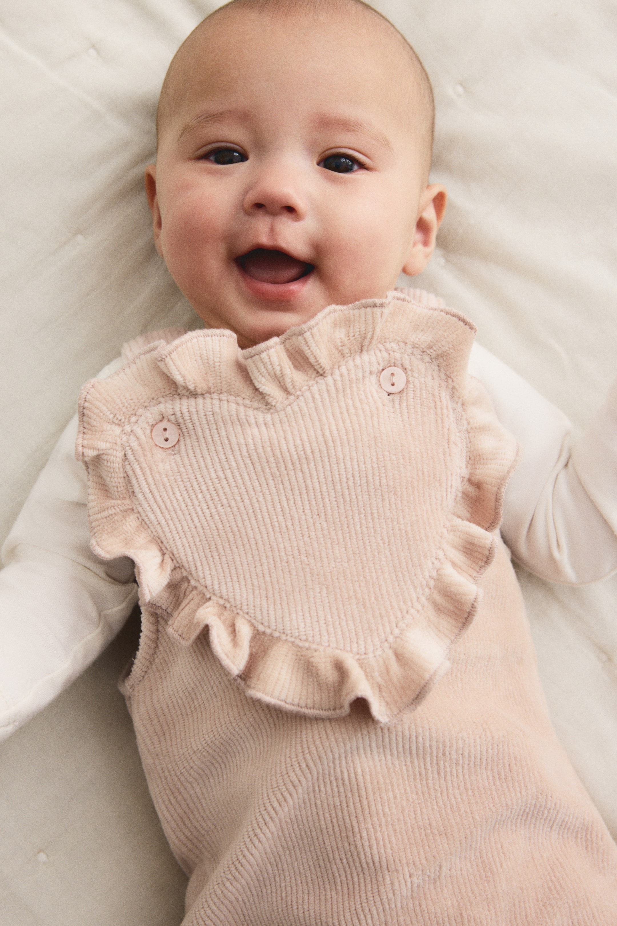 View larger image: A light pink corduroy overall is featuring a heart-shaped bib with ruffled edges and two decorative buttons. It is styled over a white long-sleeved top and is presented from a top-down perspective.