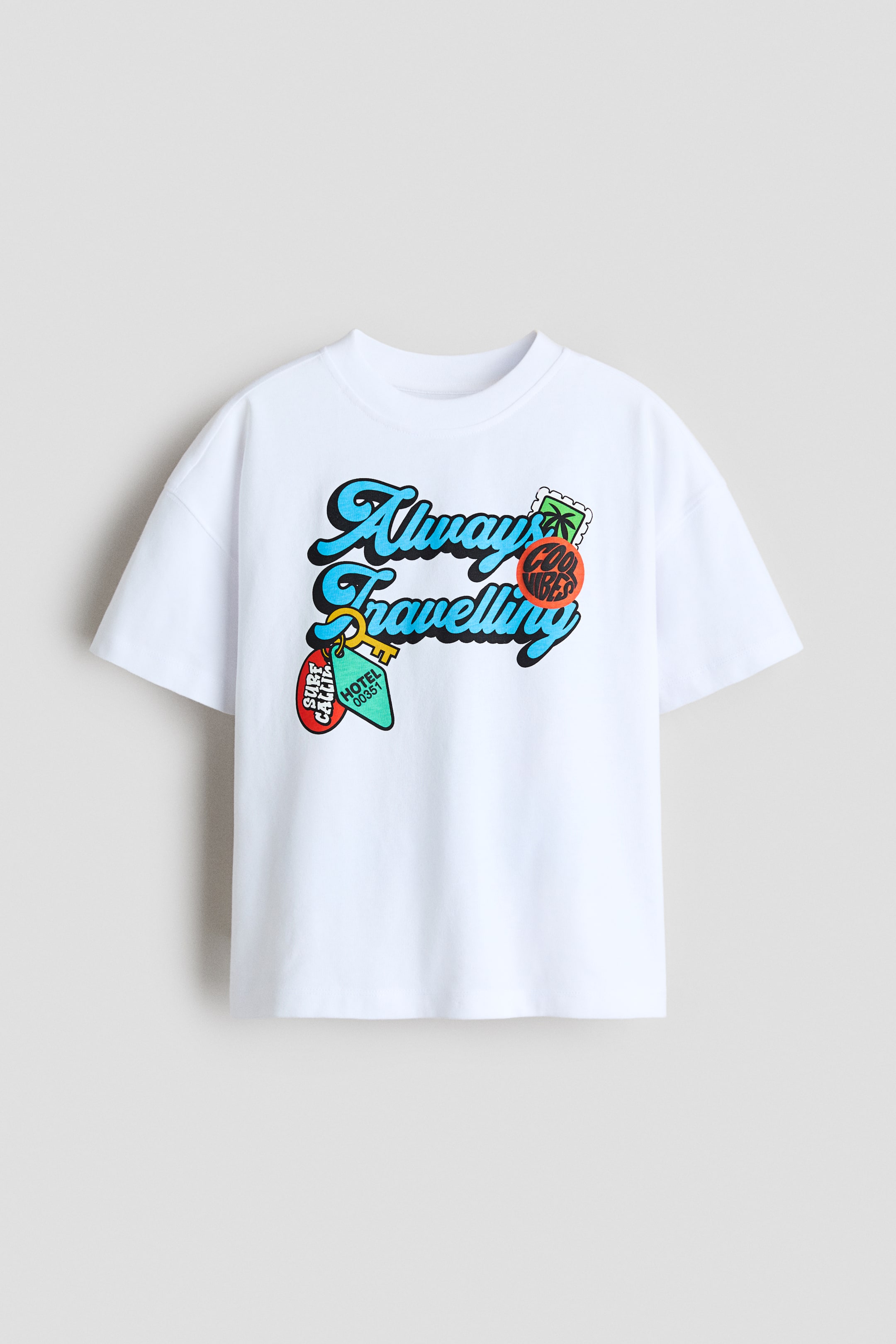 Agrandir l'image: Un t-shirt blanc à coupe ample en jersey de coton doux est présenté de face, avec un col rond bordé de côtes et des épaules tombantes. Le devant affiche un motif imprimé coloré avec le texte "Always Travelling", un palmier, un timbre et des étiquettes de clé.