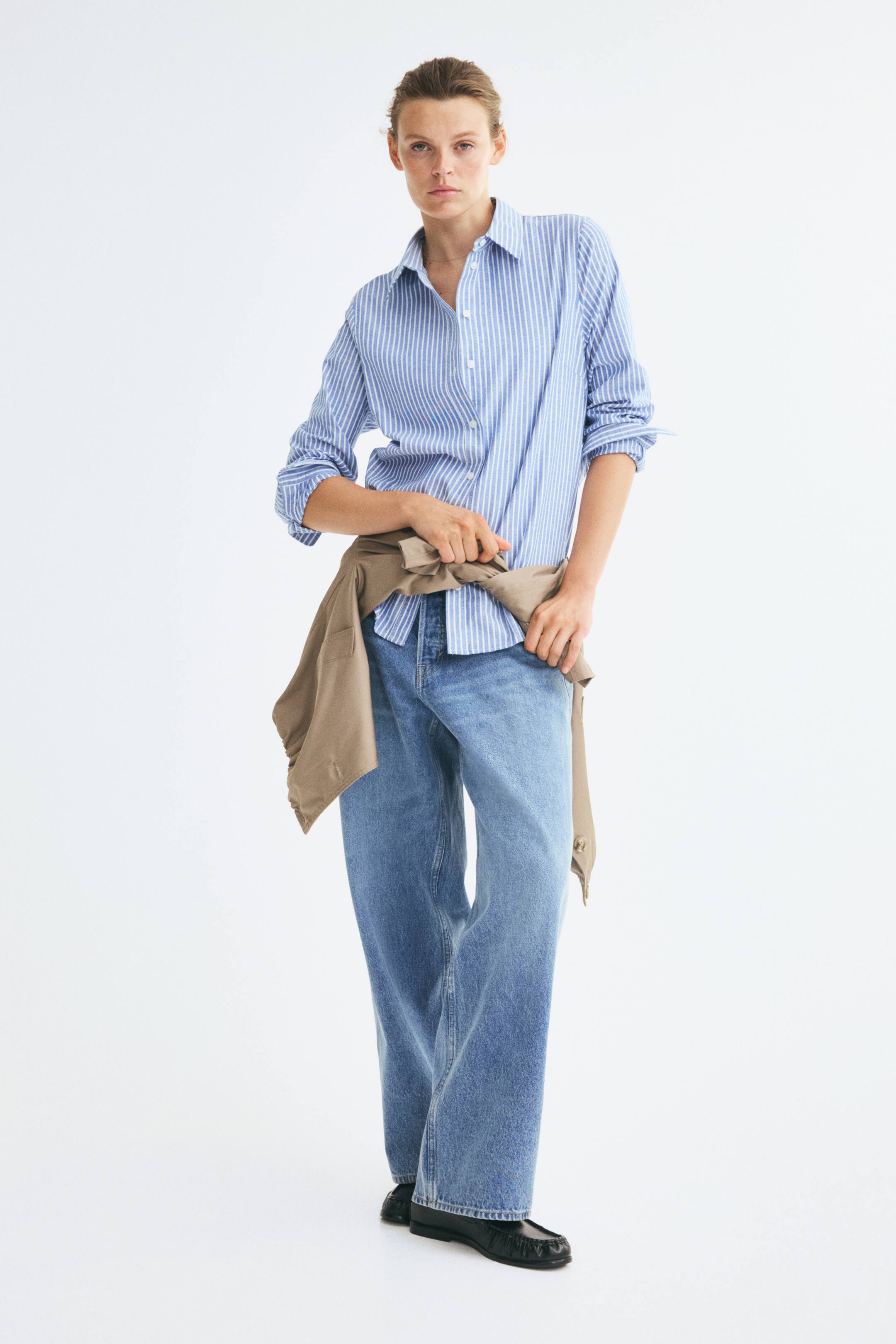 Xem ảnh lớn hơn: Một chiếc áo sơ mi Oxford cotton kẻ sọc dọc xanh lam và trắng đang được người mẫu mặc, tay áo xắn lên và cổ áo mở cúc. Áo được phối cùng quần jeans xanh ống rộng và một chiếc áo khoác màu be buộc quanh eo, được người mẫu khẽ cầm giữ.