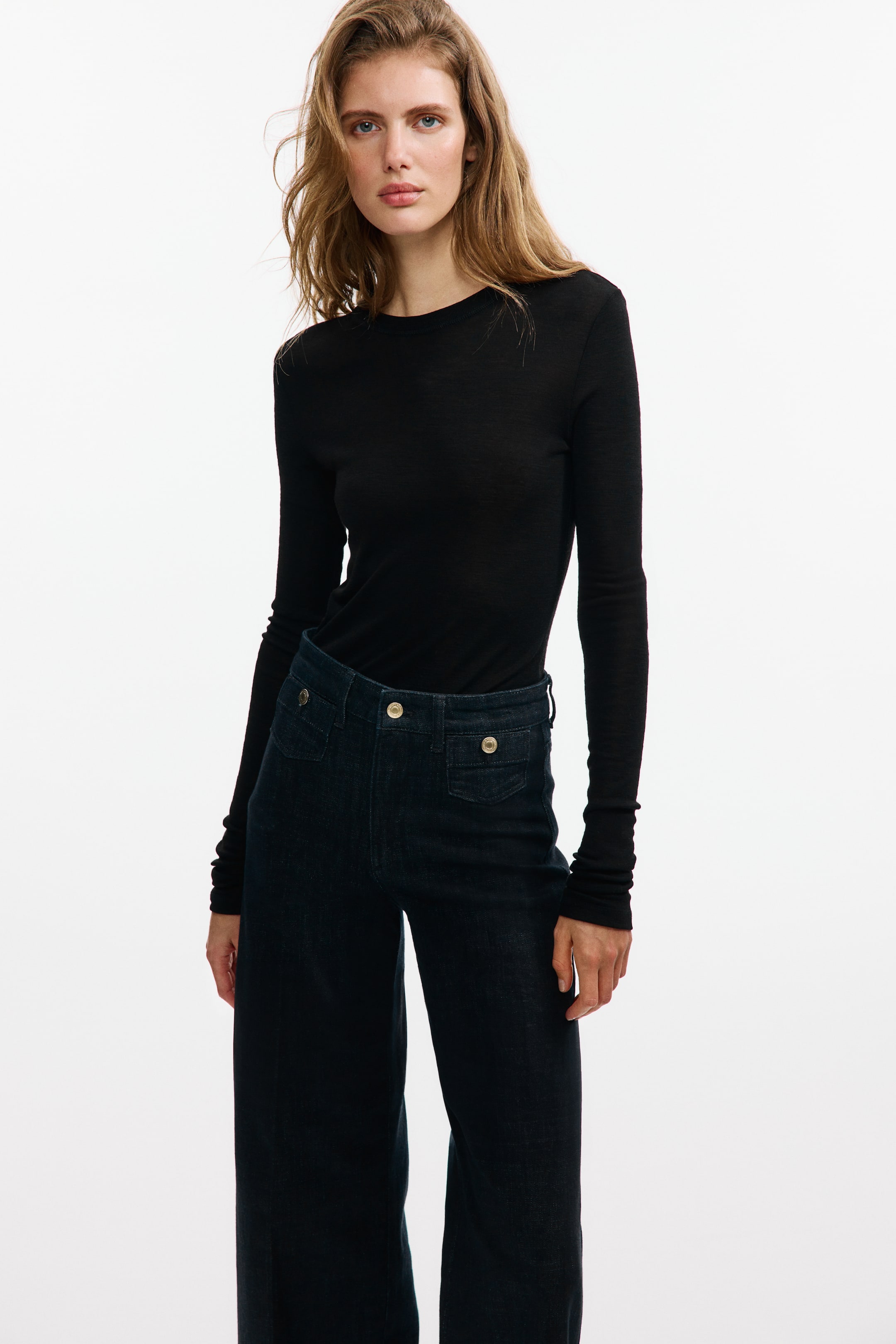 View larger image: The model is wearing dark wash denim jeans that highlight a high waist, a visible button closure, and patch front pockets with buttons. These jeans feature creases down the front and a wide-leg cut, fitting slim from the waist to the hip. A black long-sleeved top is neatly tucked into them.