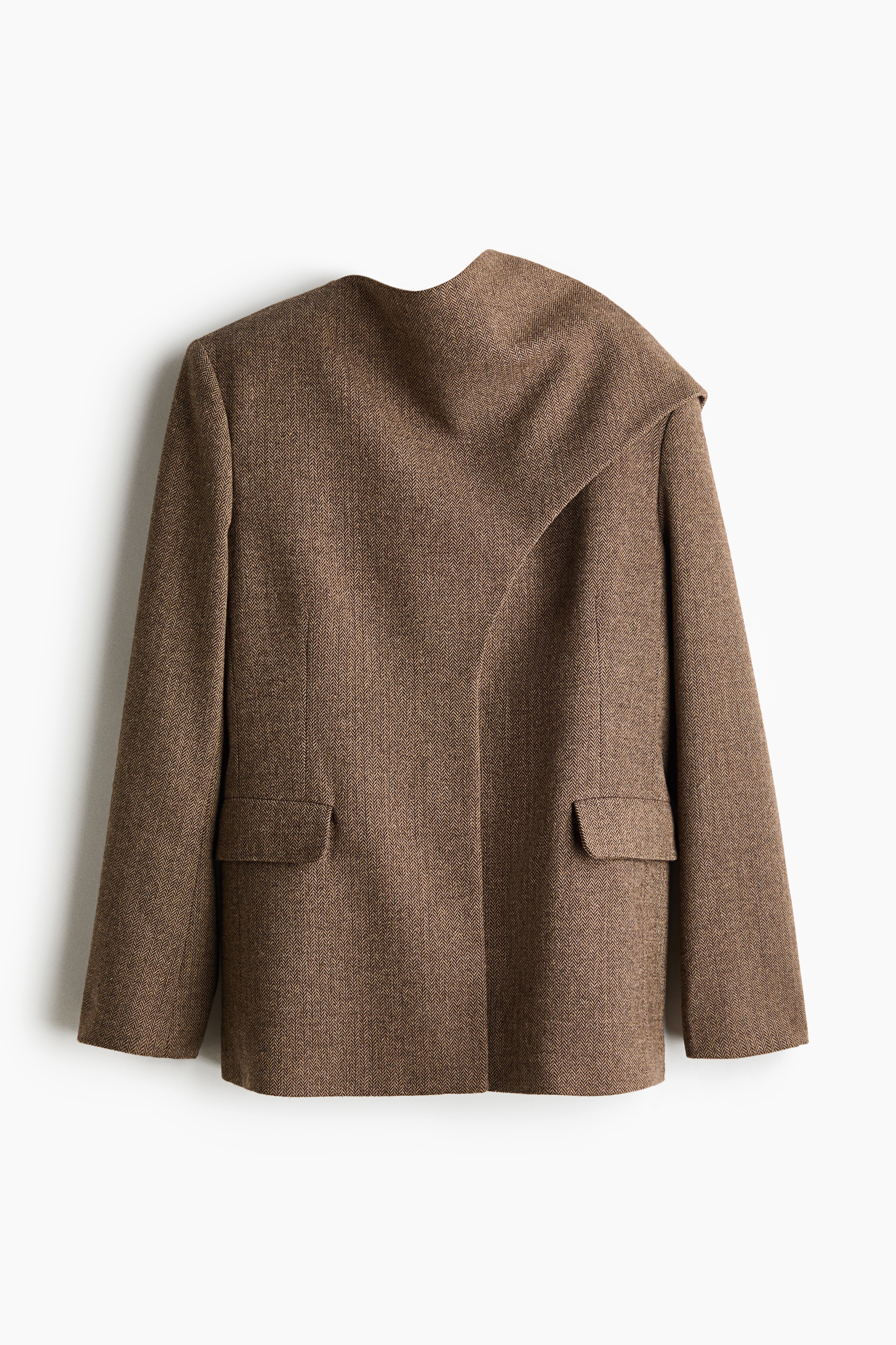 The model is wearing a brown herringbone-weave blazer, featuring structured shoulders and a front panel draped over one shoulder as a scarf-like detail. The blazer is styled over a white roll-neck top and paired with wide-leg blue jeans.