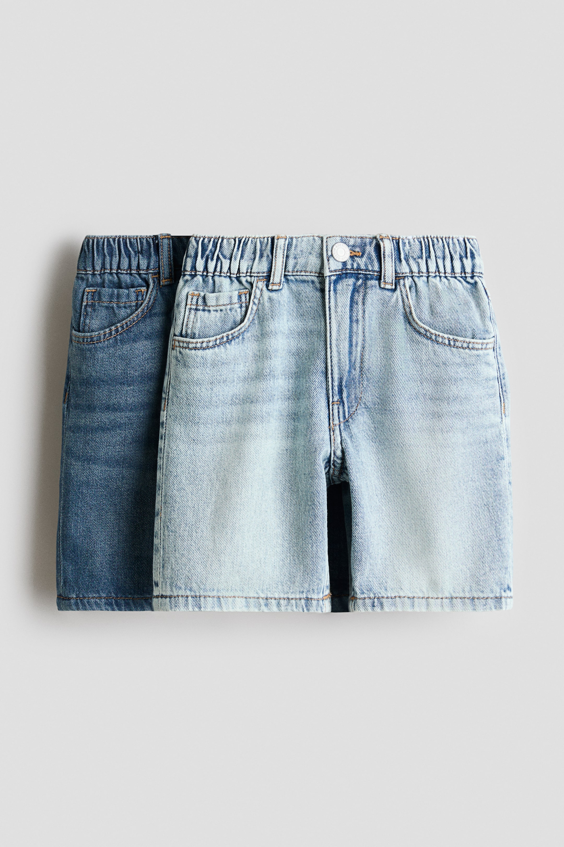 View larger image: Two pairs of knee-length, 5-pocket shorts are showcasing their front view, with a light wash denim pair prominently in the foreground and a darker wash denim pair positioned slightly behind. Both shorts feature an elasticated waist and front pockets, while the front pair also displays a press-stud closure accompanied by a zip fly.