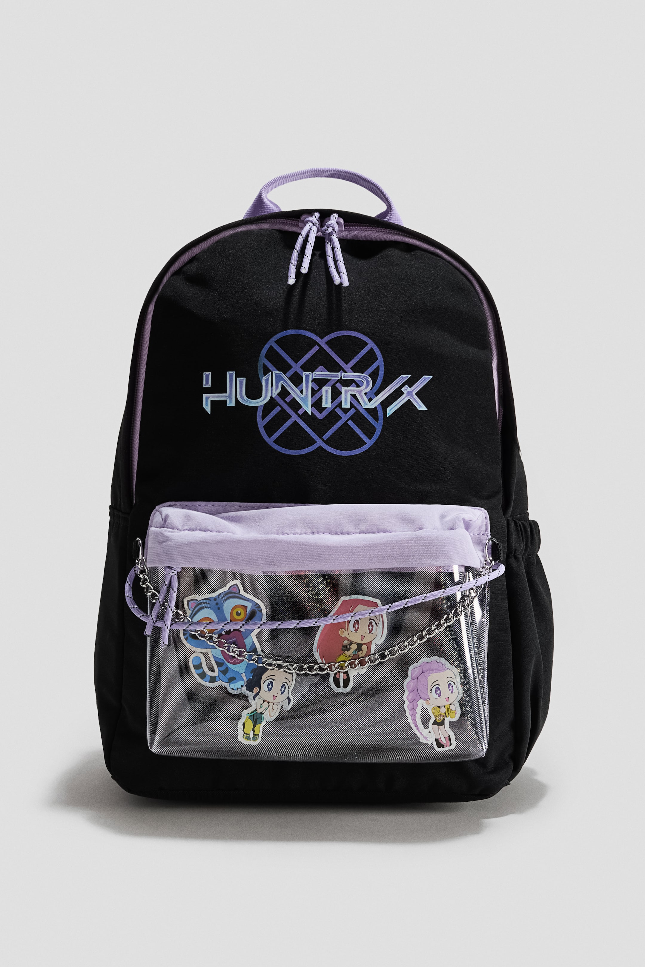 View larger image: The black backpack is facing front, featuring a holographic "HUNTRX" logo on its main compartment and purple details including a top handle and zipper pulls. A clear PVC front pocket displays four cartoon character patches, with a silver-toned chain and purple rope draped across it, and a purple drawstring closure.